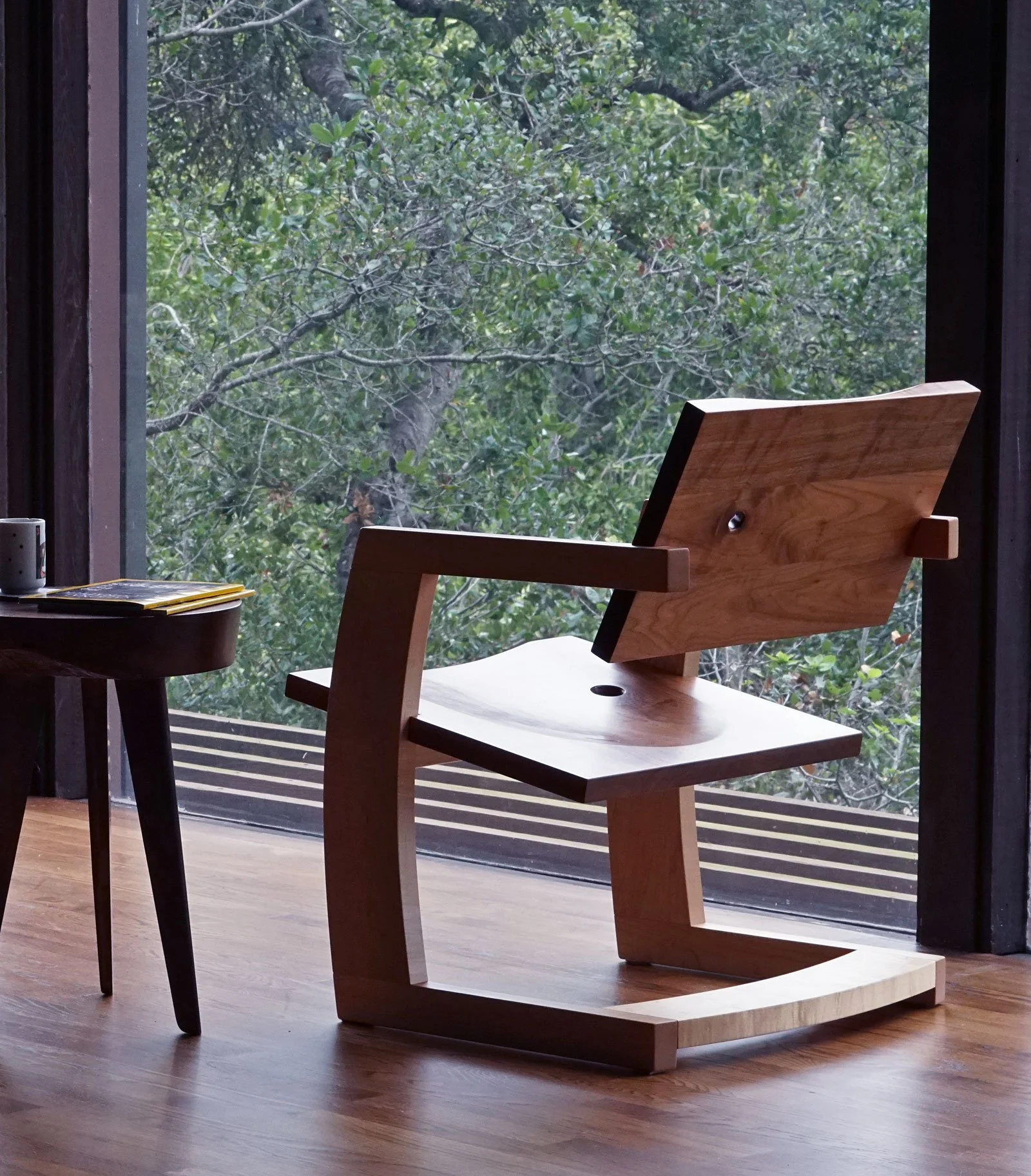 Interior scene with a wooden chair facing a large window showing green leafy trees outside, a small black side table with a decorative mug and some magazines on it.