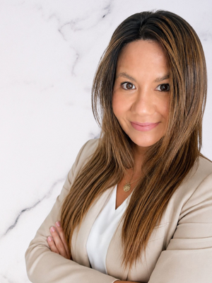 A woman with long brown hair and light makeup, wearing a beige blazer and white top, standing against a white marbled background.