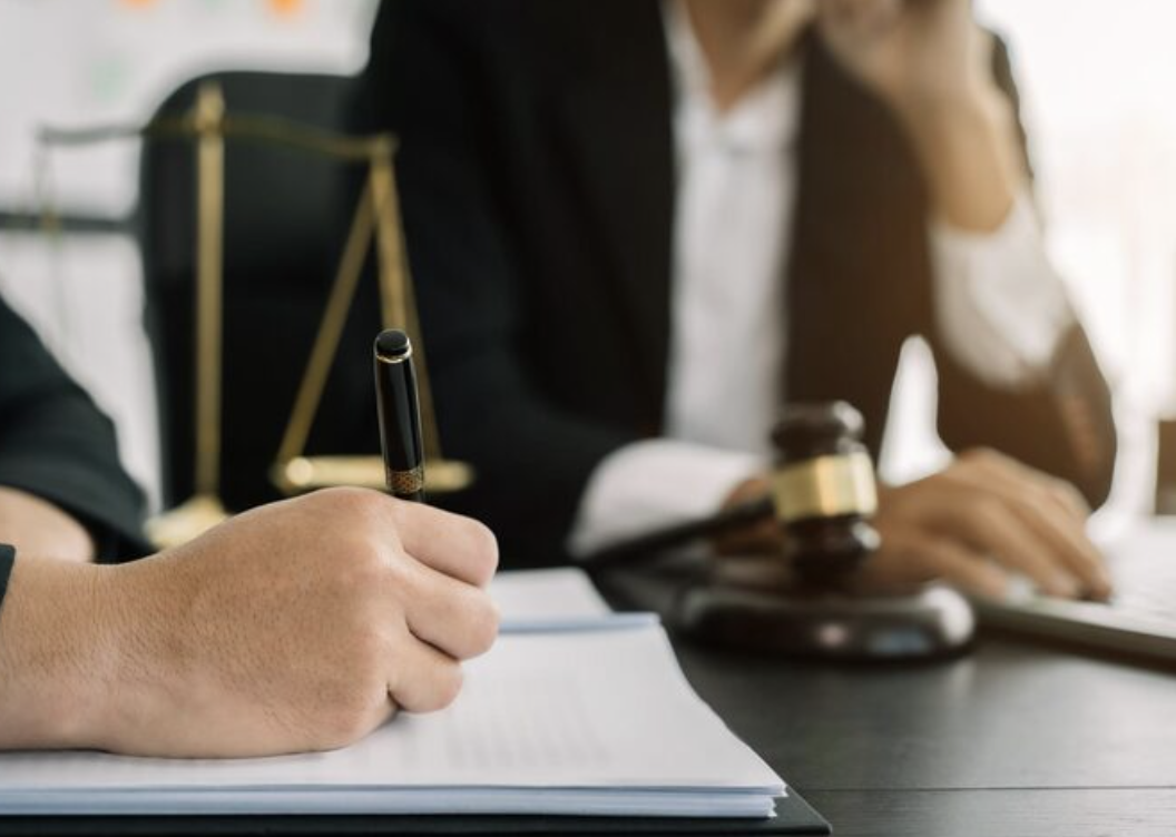 A person signing a document with a pen, with a judge's gavel and scale of justice in the background.