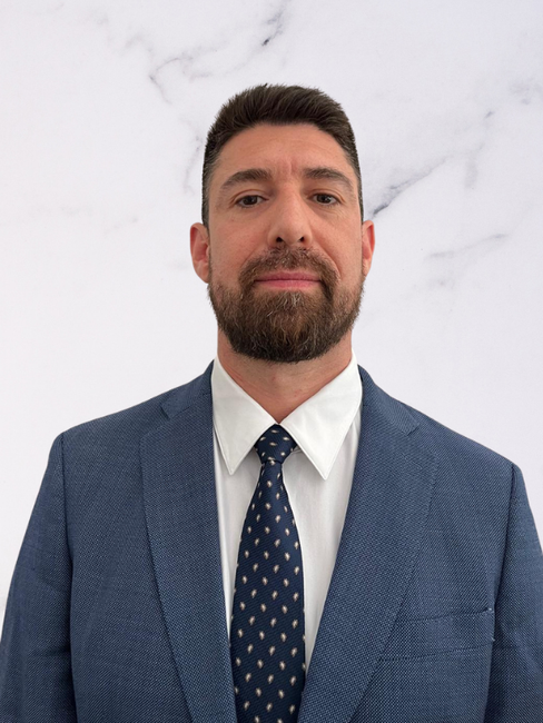 Man with dark hair, beard, and mustache wearing a blue suit, white shirt, and dark patterned tie, standing against a light gray marble background.