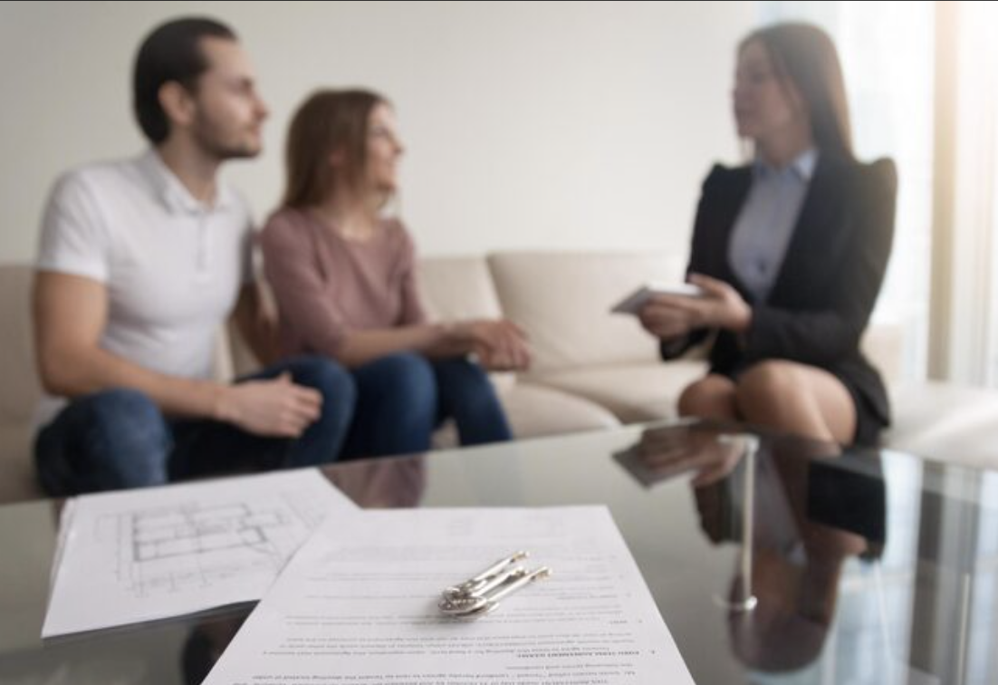 Three people sitting on a sofa during an interview or consultation with a female professional holding a notepad, and a glass table in the foreground with documents and keys.