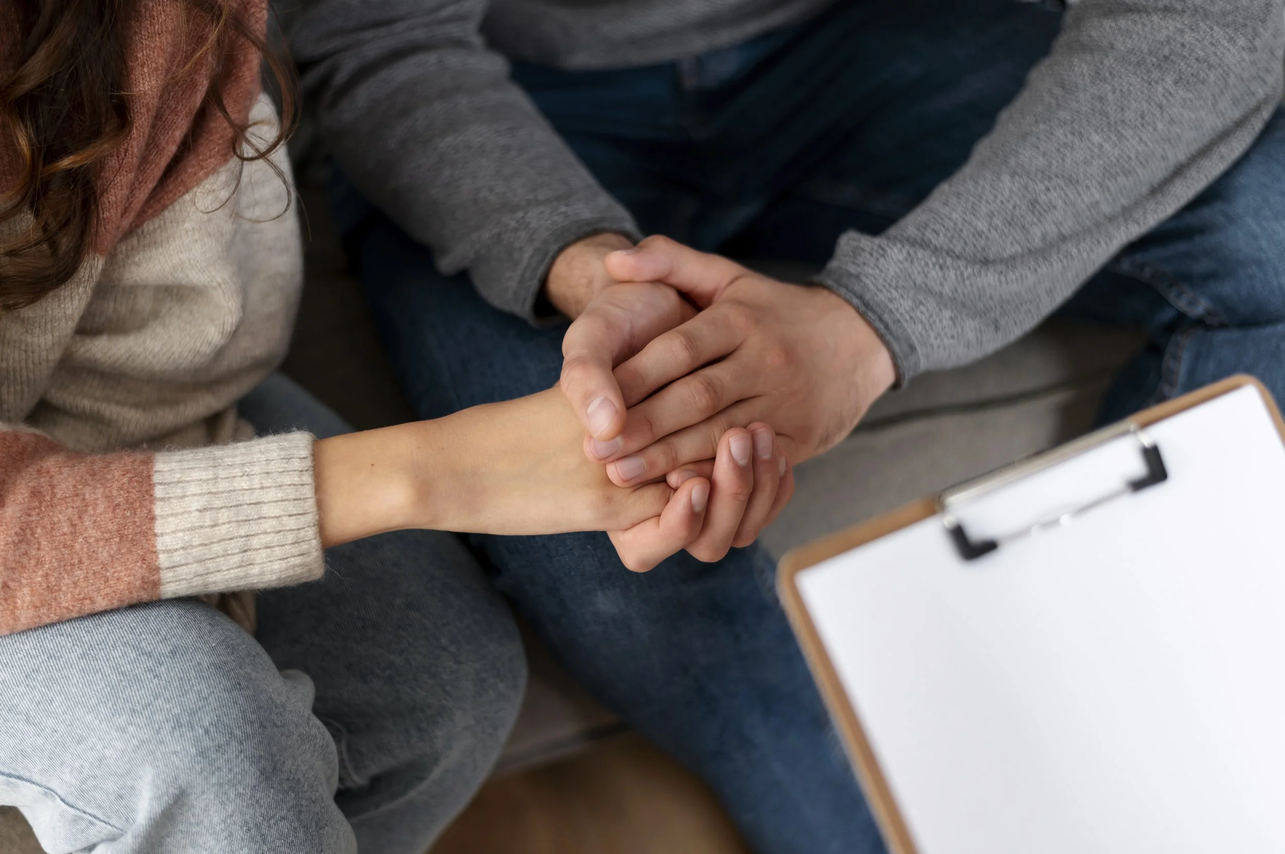 A woman and a man holding hands, with the women wearing a cream and peach sweater and the man wearing a gray sweatshirt, sitting next to each other.