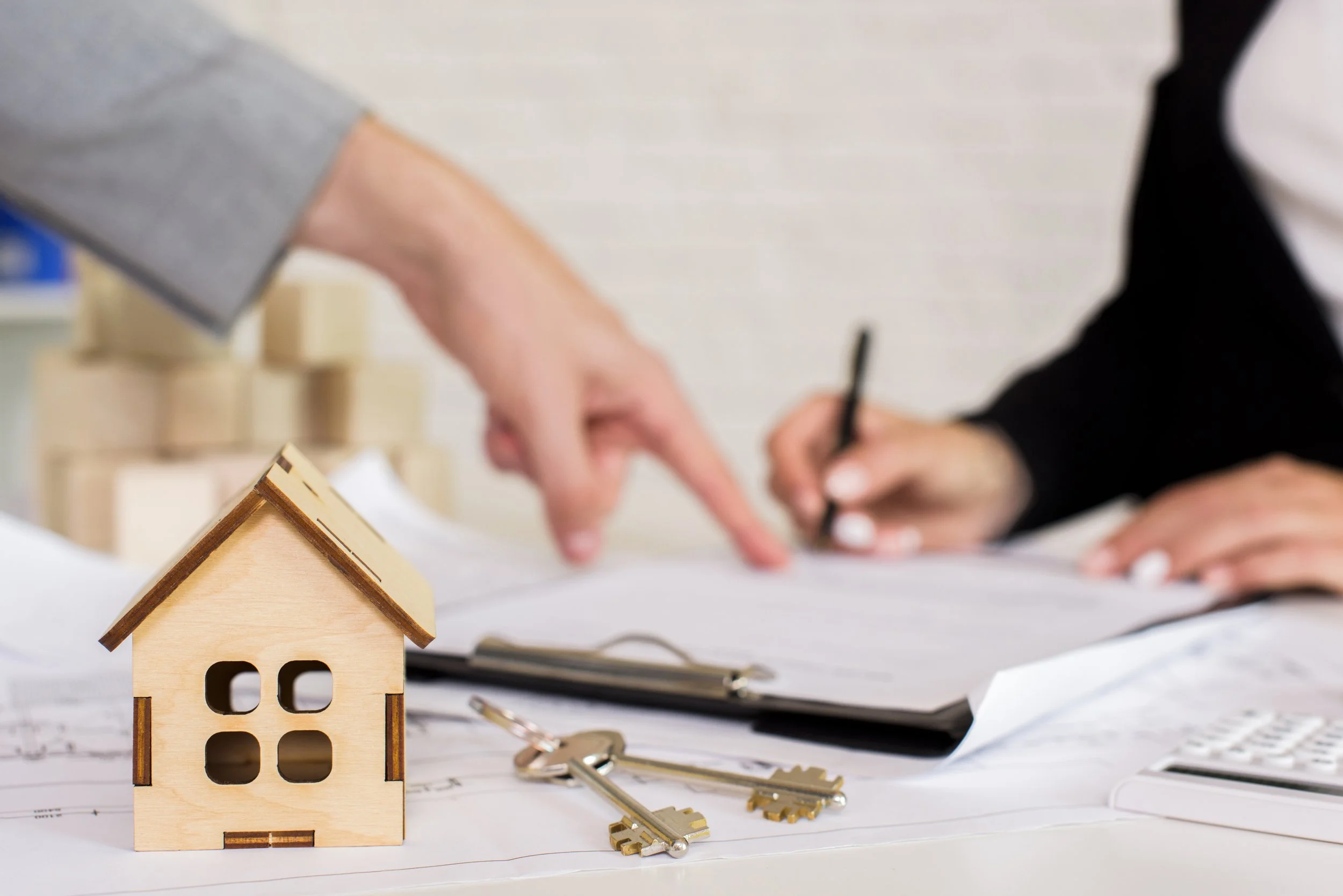 Close-up of a small wooden house model on paper with a set of keys and blurred hands in background, likely at an office or real estate setting.