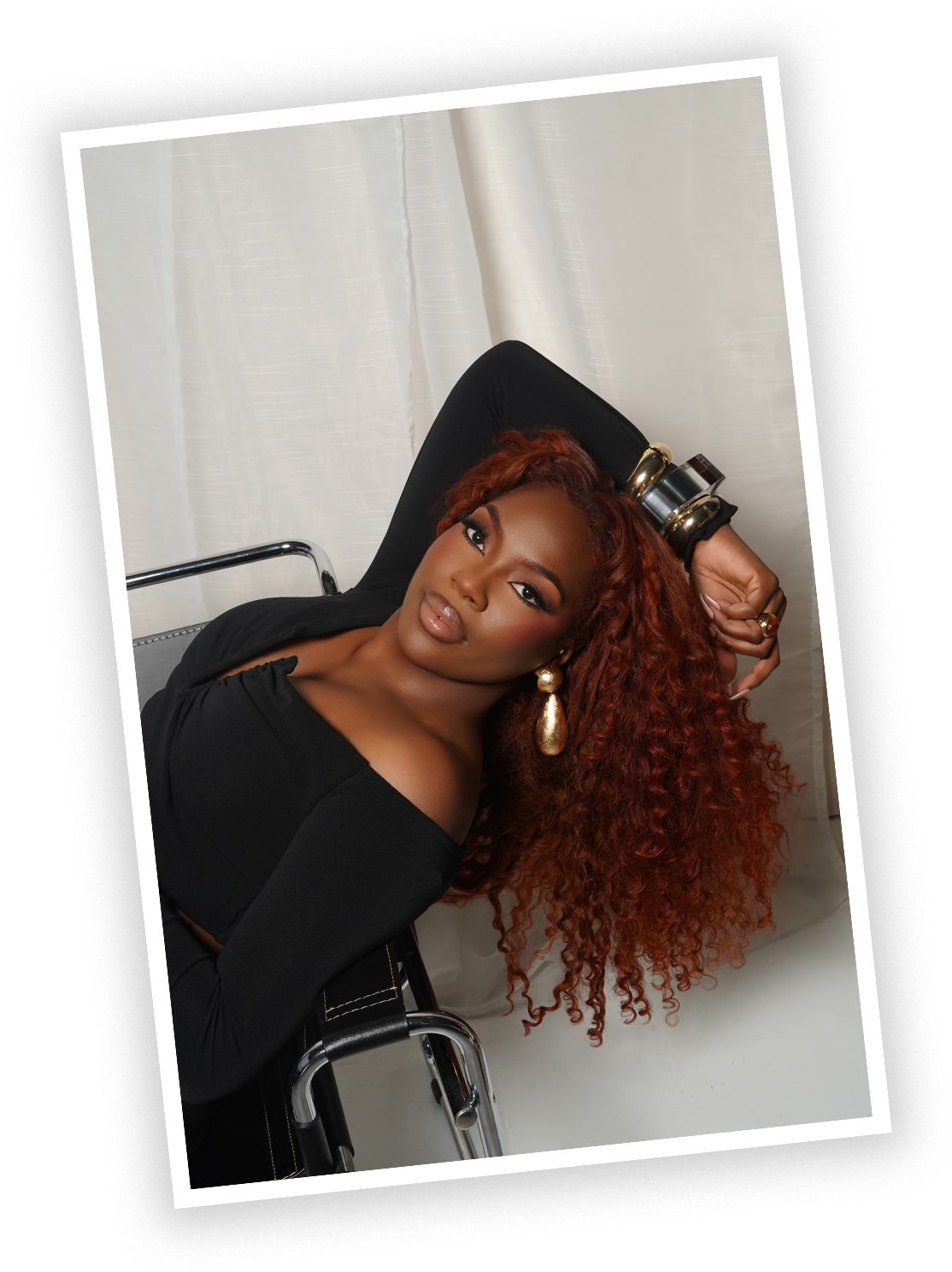 A woman with curly red hair sitting in a black chair with white curtains in the background, wearing a black top and large gold earrings.