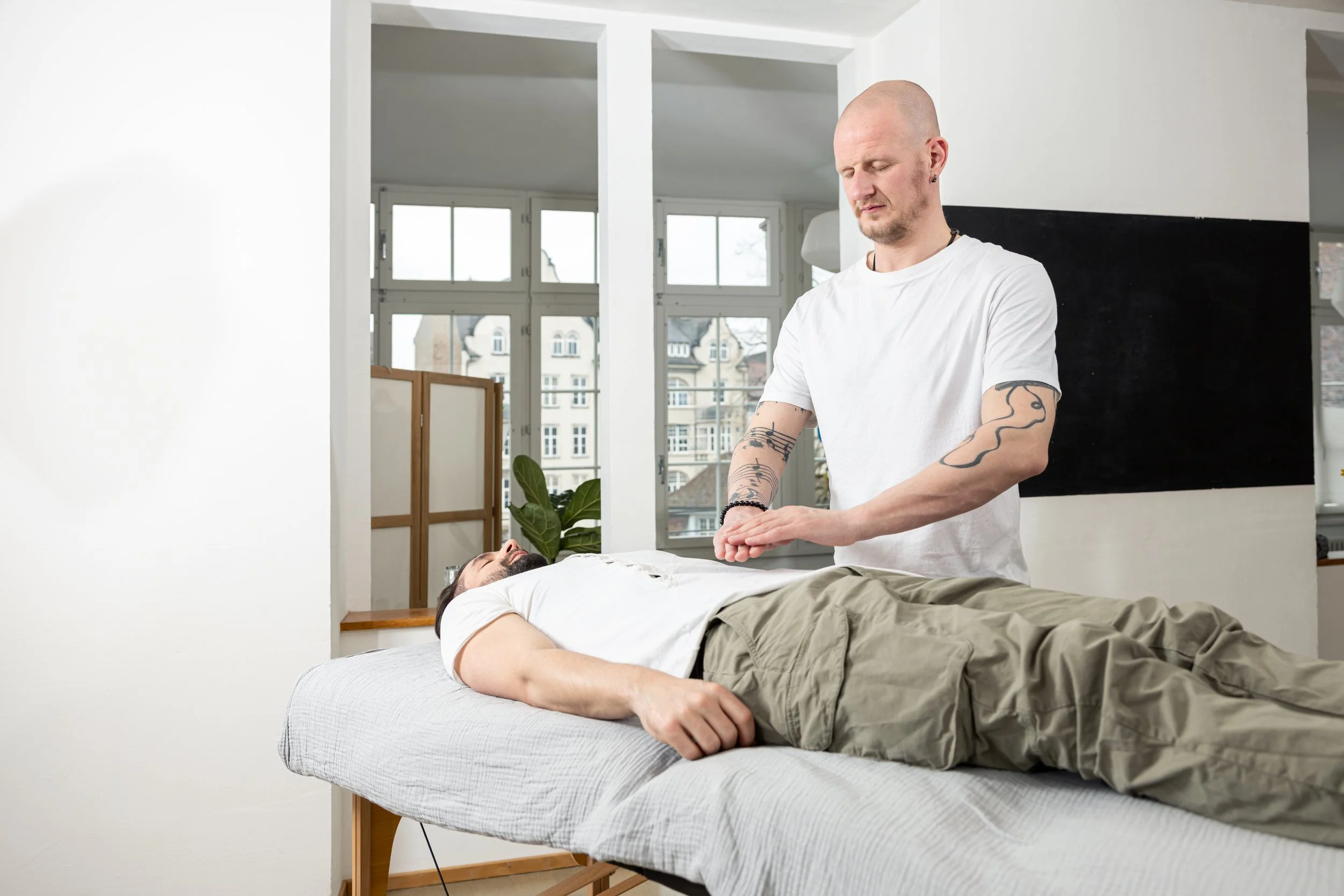 Ein Mann liegt auf einer Couch mit geschlossenen Augen, während eine tätowierte Frau neben ihm steht und seine Hand hält, in einem hellen Raum mit großen Fenstern.