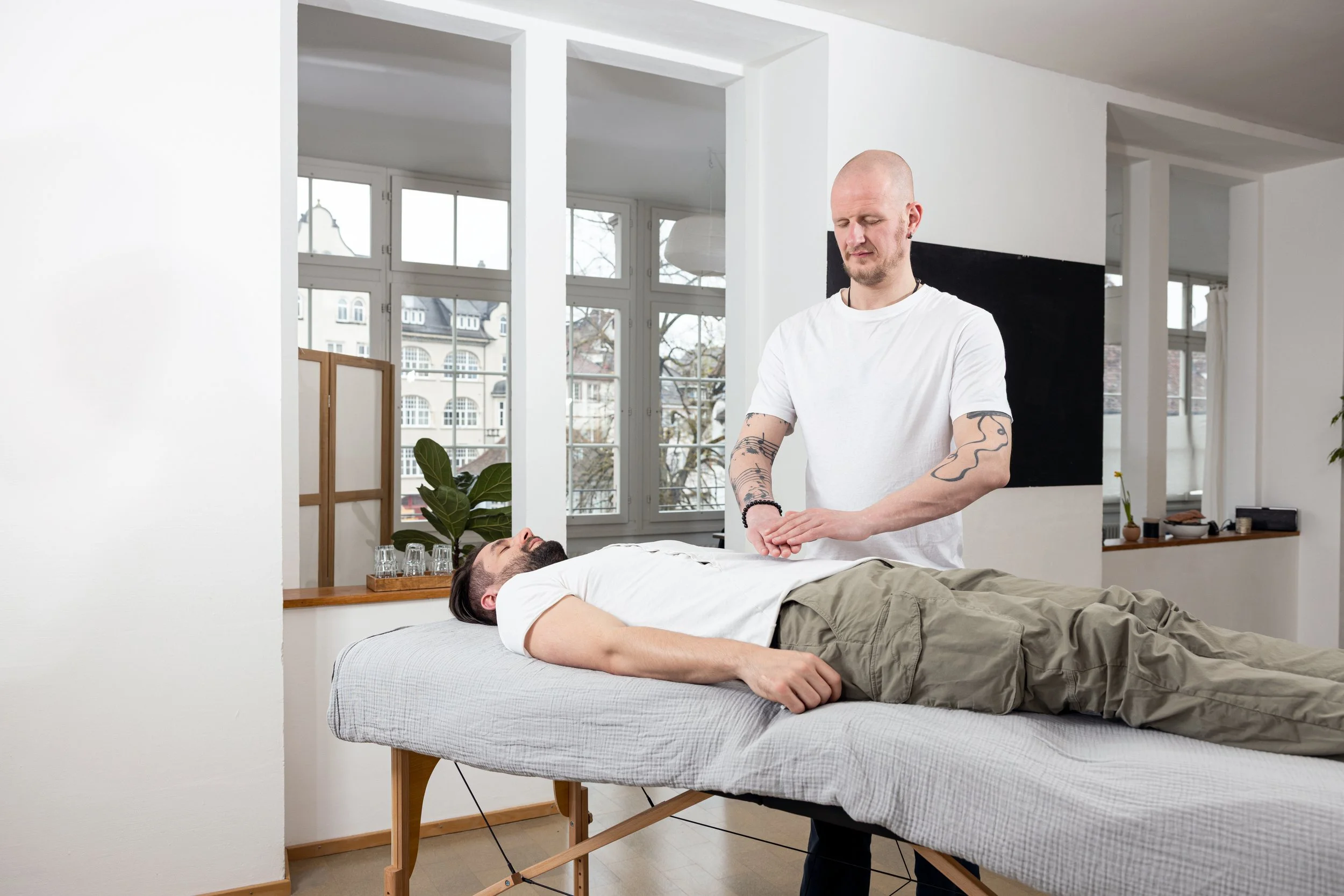 Mann liegt auf einer Massageliege, während ein Therapeut ihn massiert in einem modernen Raum mit großen Fenstern und winterlicher Aussicht.