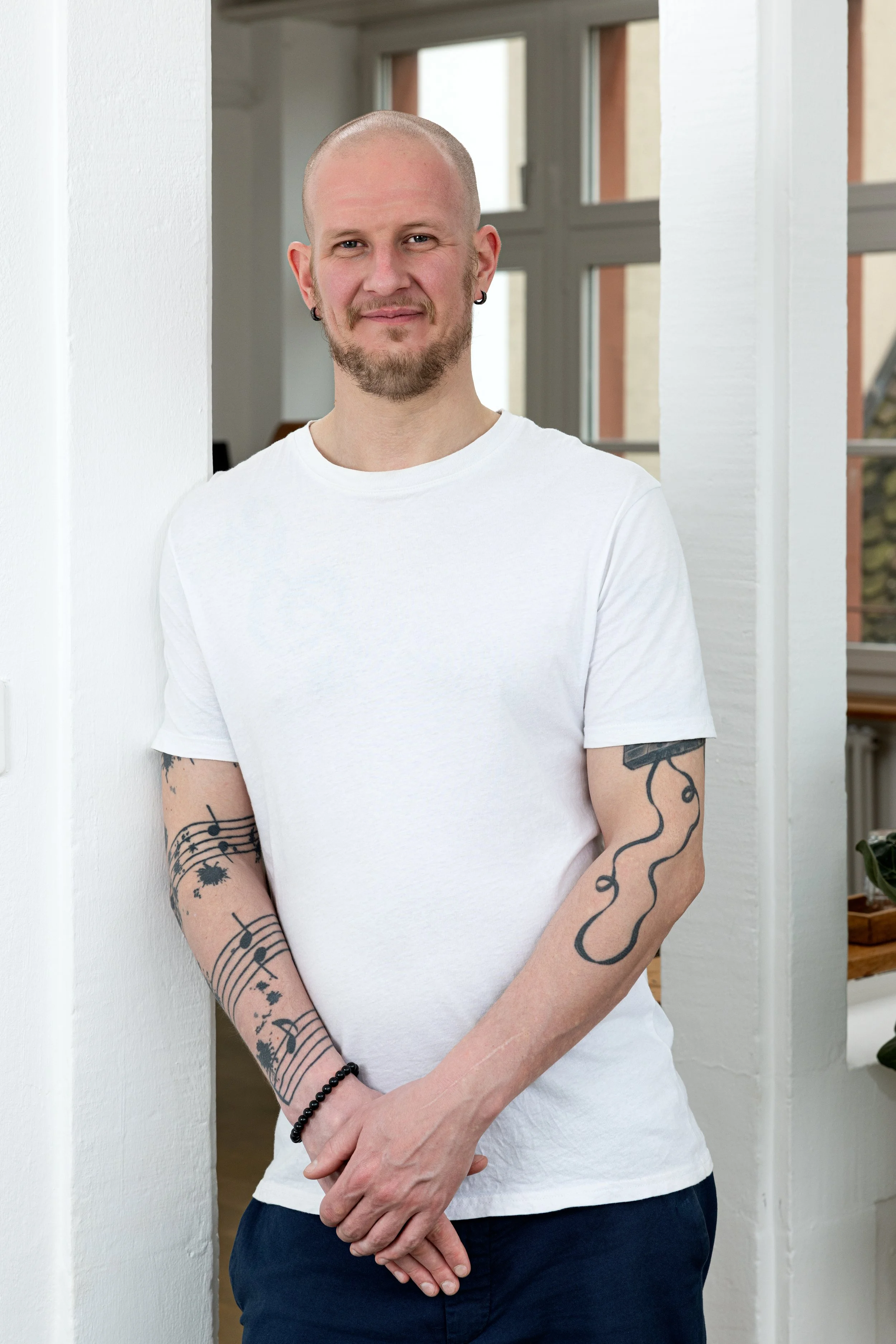 Junge Mann mit Tattoos an den Armen, weißes T-Shirt, schwarze Armbanduhr, kurze hellblonde Haare, im Hintergrund weiße Wände und große Fenster