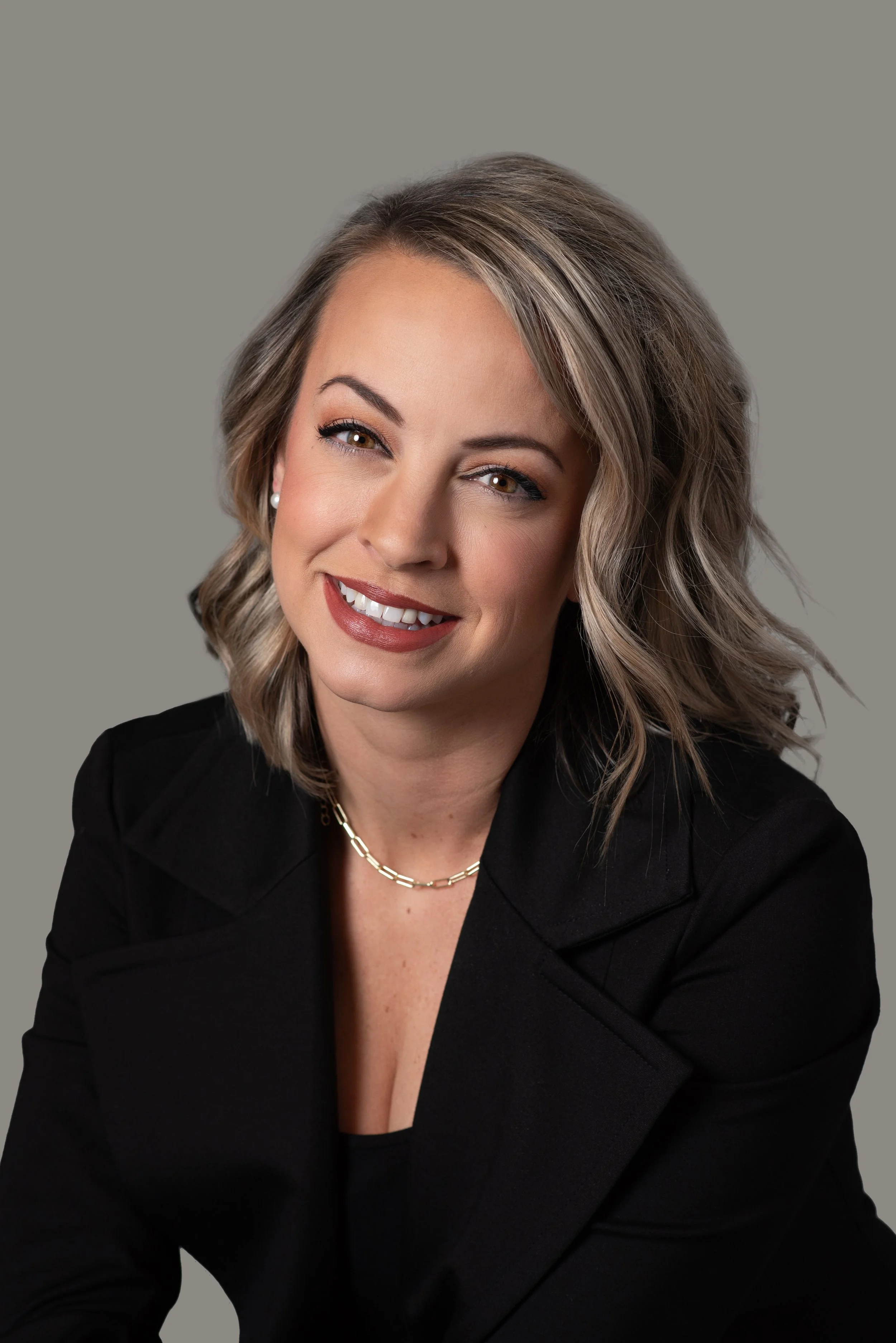 A woman with shoulder-length blond hair, smiling with white teeth, wearing a black blazer, pearl earrings, and a gold chain necklace, set against a gray background.