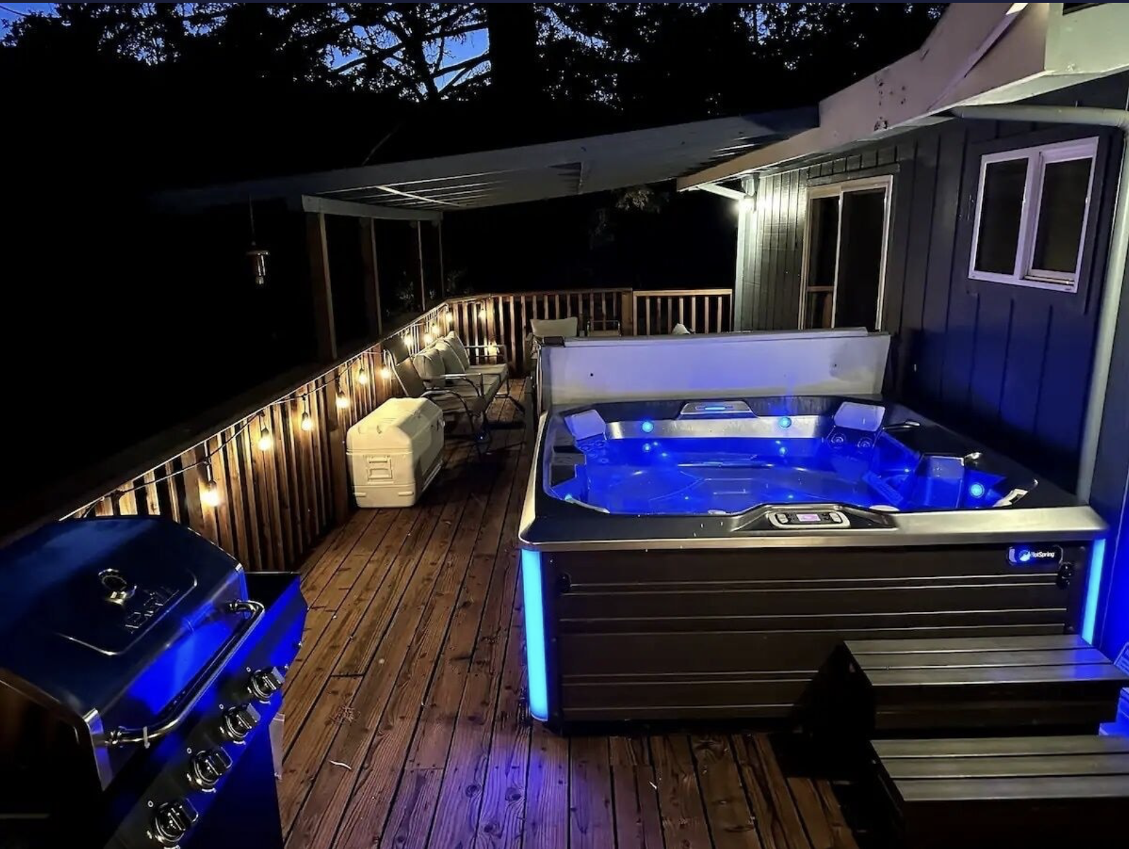A nighttime outdoor deck with string lights, outdoor seating, a hot tub with blue lights, and a grill.