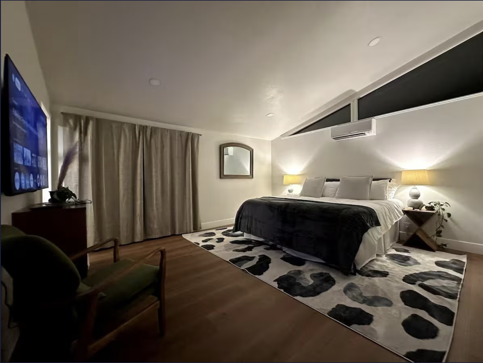 Cozy bedroom with a large bed, nightstands with lamps, a wall mirror, a mounted flat-screen TV, curtains covering a window, and a patterned rug on the wooden floor.