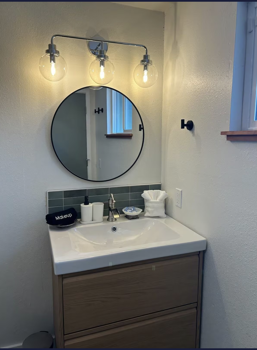 Bathroom vanity with oval mirror, three-light fixture, and wall-mounted towel hook. Contains a sink with soap, lotion, and tissue, with a small window above.