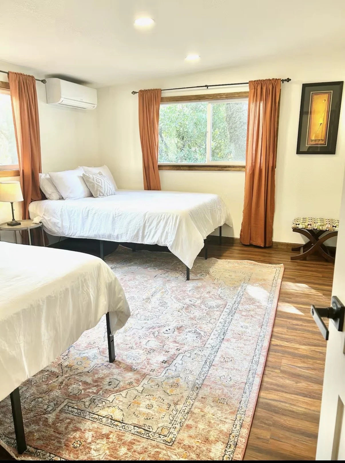 A cozy bedroom with two beds, white bedding, and several pillows. The room has wooden floors, a window with orange curtains, and a patterned rug. There is a small table with a lamp, wall art, a bench near the window, and an air conditioning unit above the window.