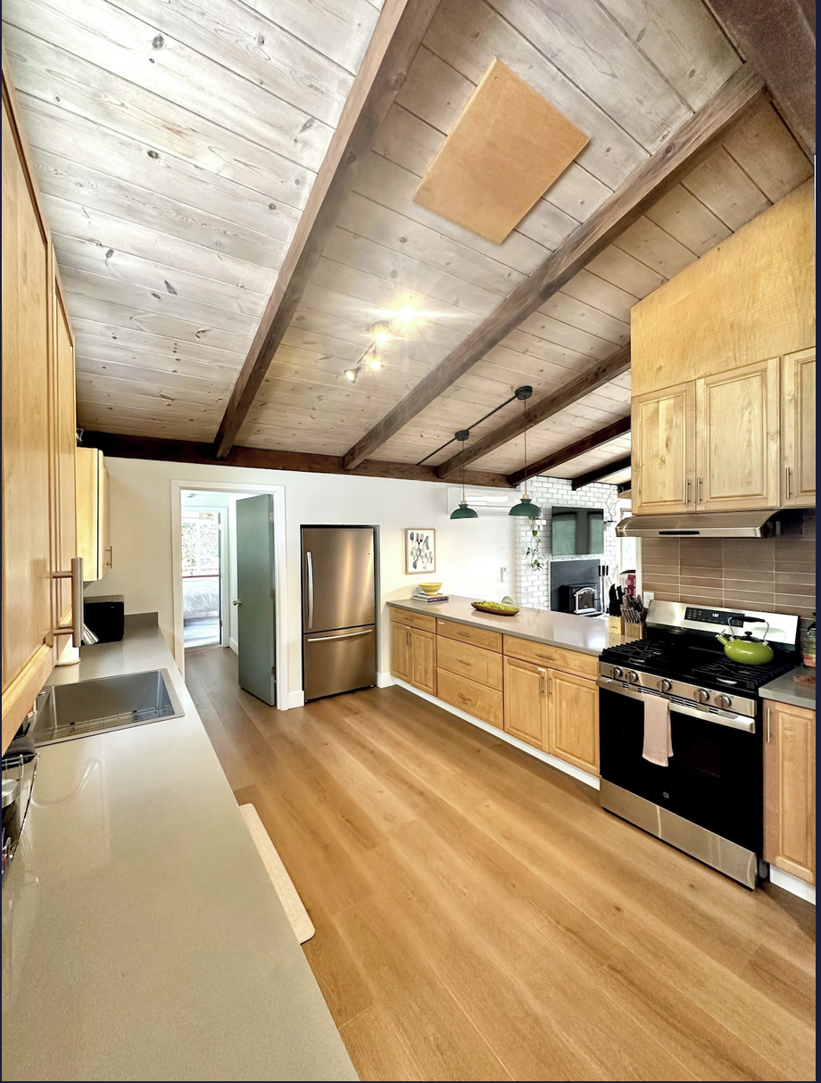 Kitchen with wooden cabinets, stainless steel appliances, a white countertop, wood flooring, pendant lights, and a vaulted ceiling with wooden beams.