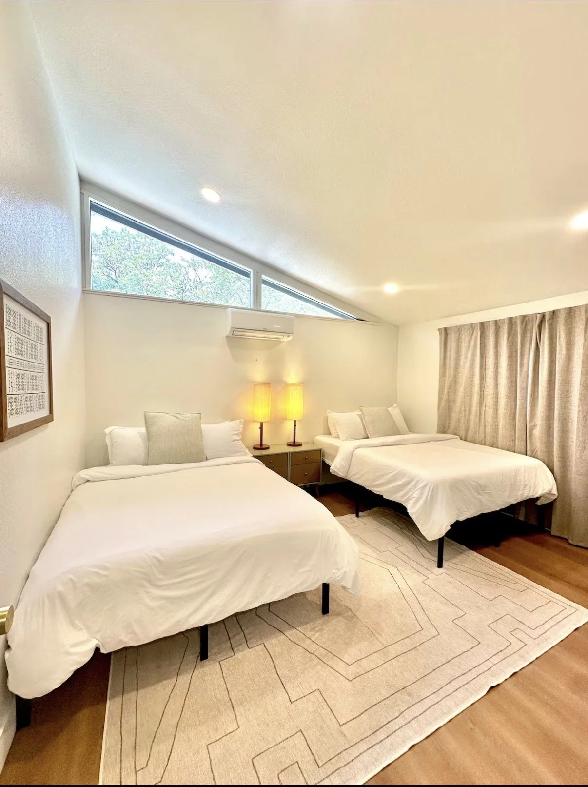 A bedroom with two beds, a large window, beige curtains, a beige rug, and two lamps on a nightstand.