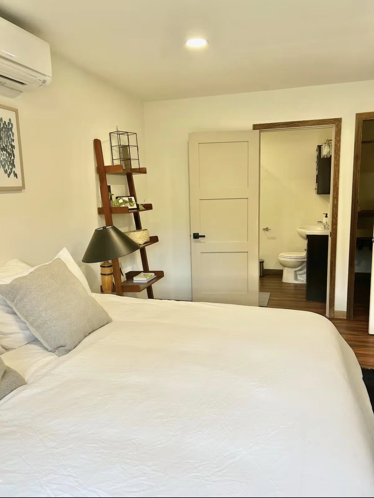 A bedroom with a white bed, beige pillows, a black bedside lamp, a wooden ladder shelf, and a small bathroom with a toilet and sink visible through a door.