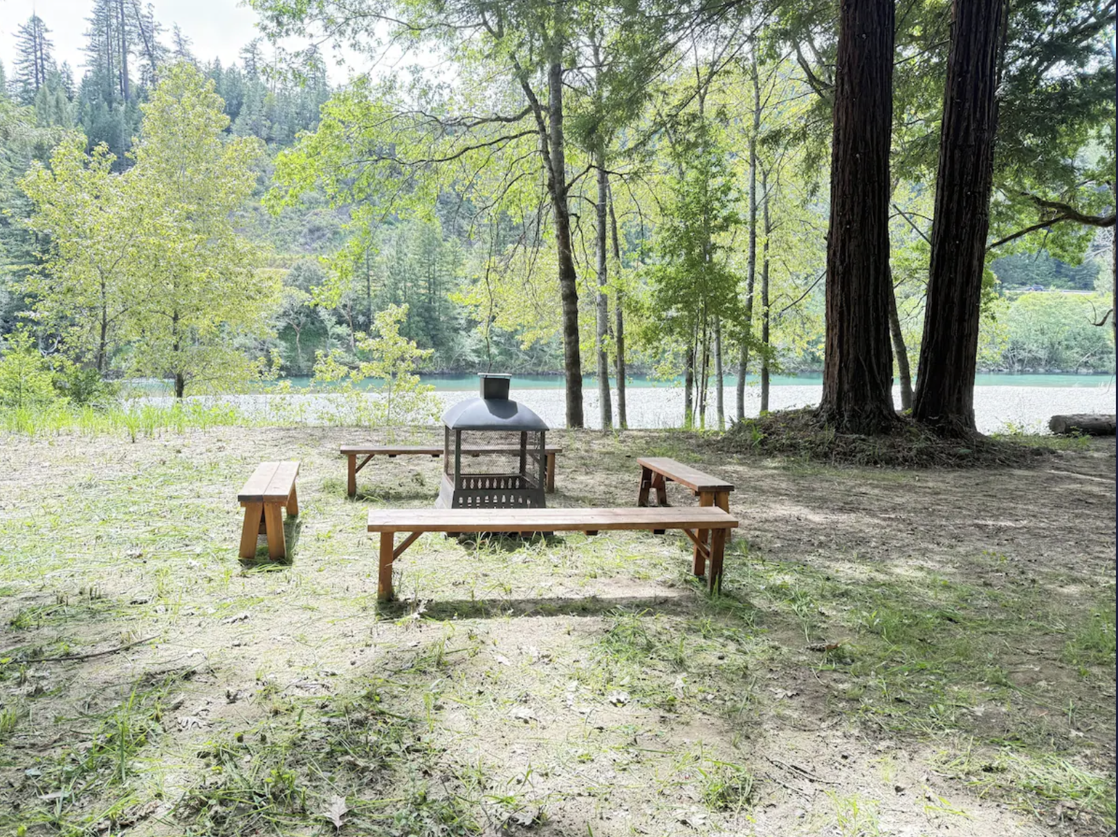 A lakeside scene with a small wooden picnic table and benches, a fire pit, tall trees with green leaves, and a calm lake in the background surrounded by lush forest.