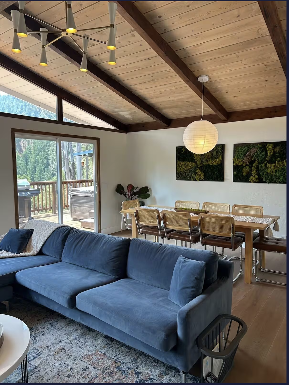 Interior of a living and dining area with large windows showing a view of trees. The room has a blue couch, a wooden dining table with seven chairs, a hanging paper lantern, and wall art consisting of green plant arrangements.