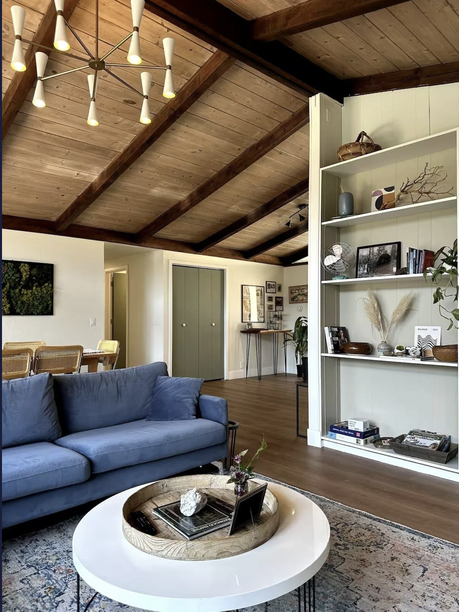 Living room with a wooden vaulted ceiling, blue sofa, white round coffee table with decor items, and a built-in white bookshelf with various decorative objects and books.