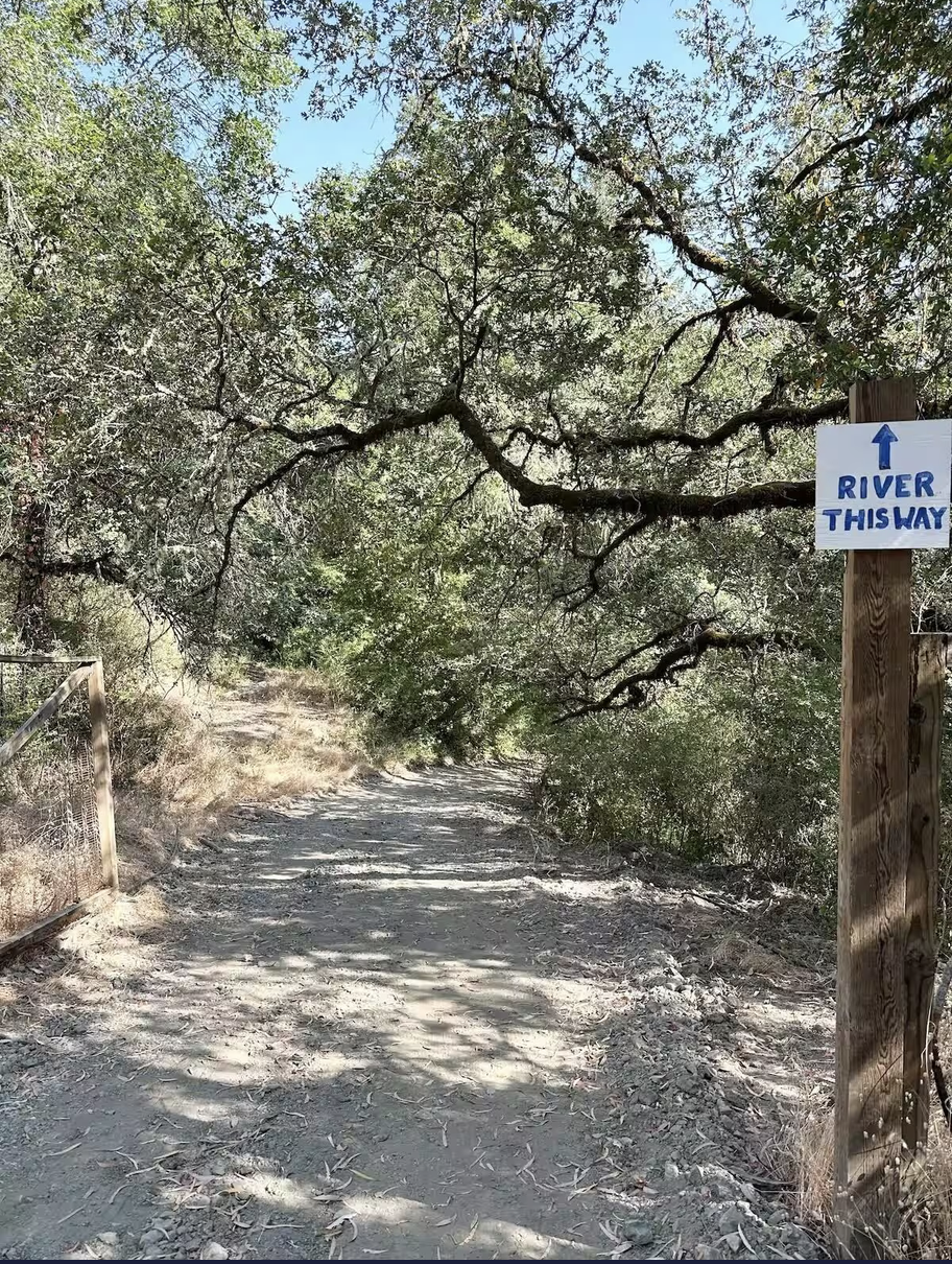 Dirt trail through woods with a sign on the right that reads 'River This Way' with an arrow pointing upward.