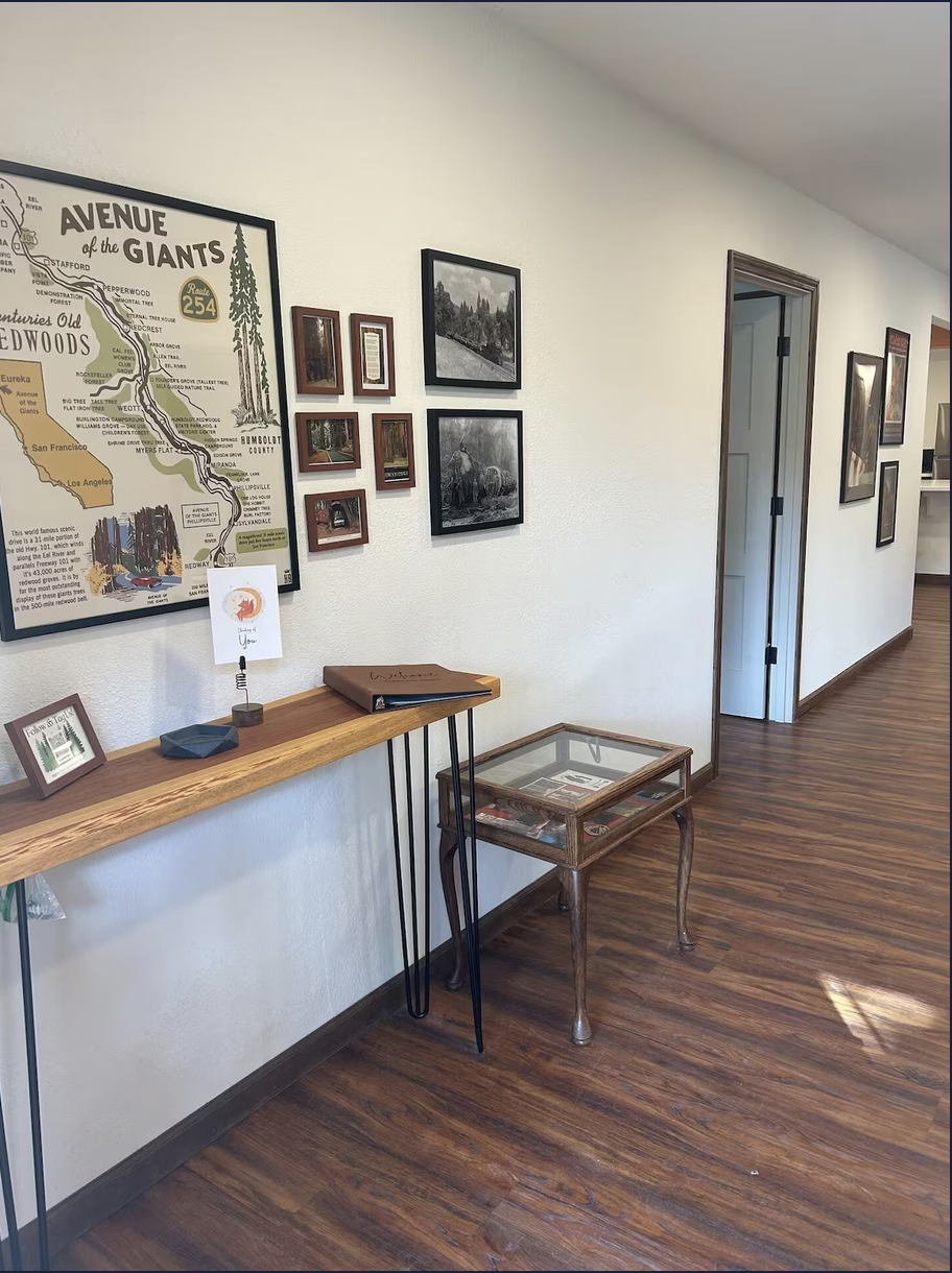 A hallway with open doorways, decorated with framed pictures and a large map on the wall. There is a wooden shelf with small items and a framed photo, and a glass-topped display table with items inside on a wooden floor.