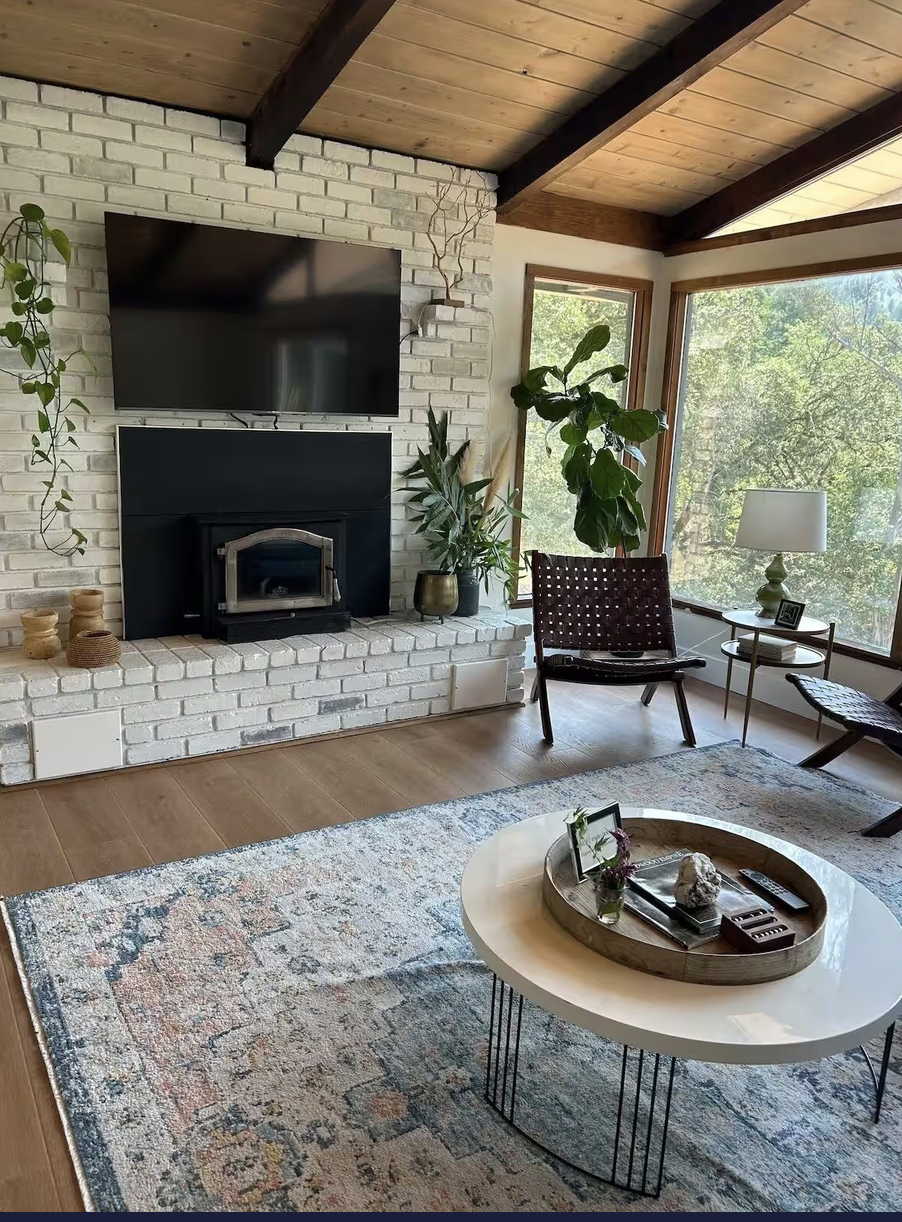 A cozy living room with large windows, a white brick fireplace, a mounted flat screen TV, a potted plant, a comfortable chair, a side table with a lamp and photo, a decorative rug, and a modern coffee table with a tray and decorative items.