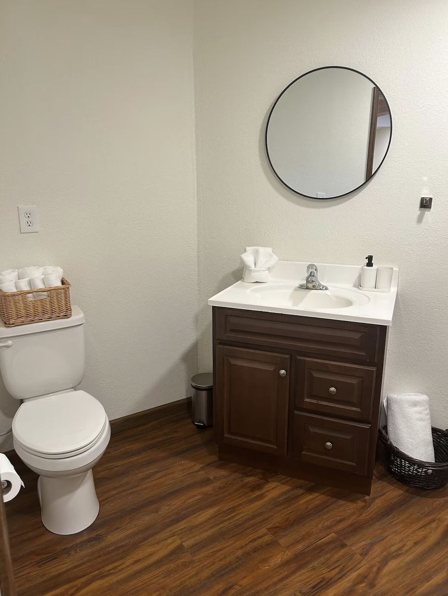 Bathroom with a toilet, wooden vanity with a white sink, round mirror, rolled towels in a basket, and a wicker trash bin with a towel inside.