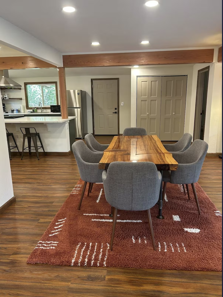 A dining room with a wooden table and six gray upholstered chairs on a red and white patterned rug. Behind, a kitchen with a refrigerator, window, and counter with bar stools.