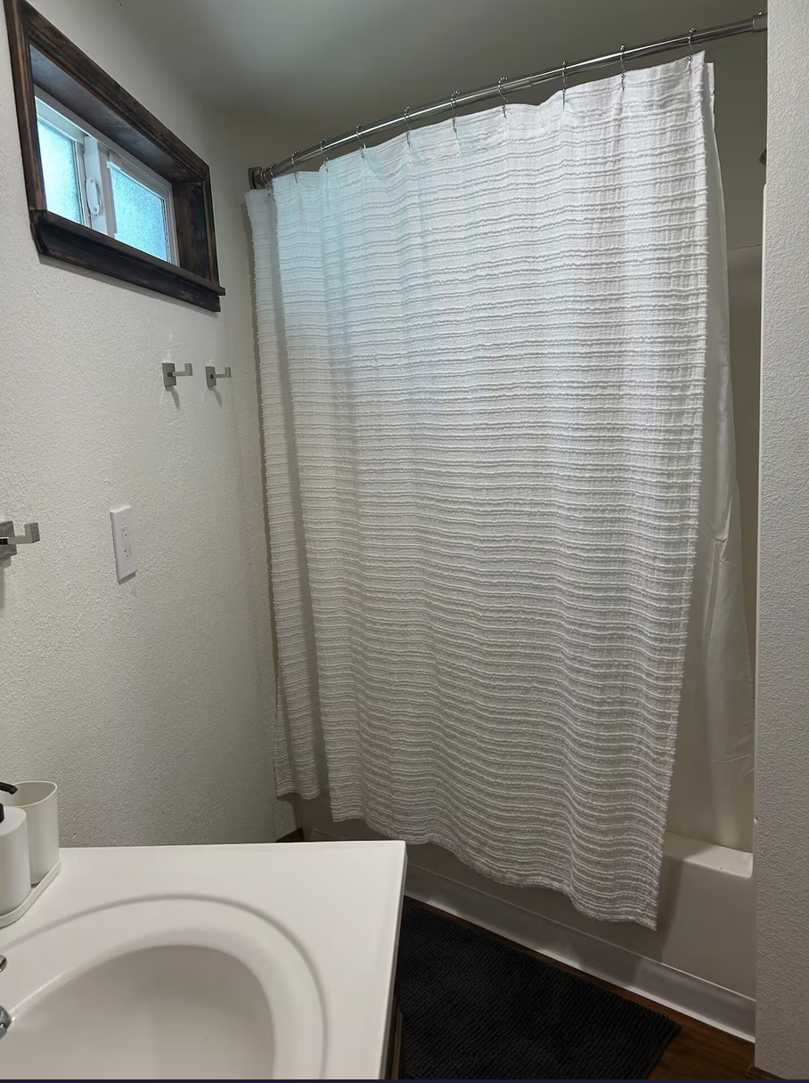 A bathroom with a small window, a mirror, a towel bar, a white shower curtain, a sink with a soap dispenser, and a dark rug on the floor.