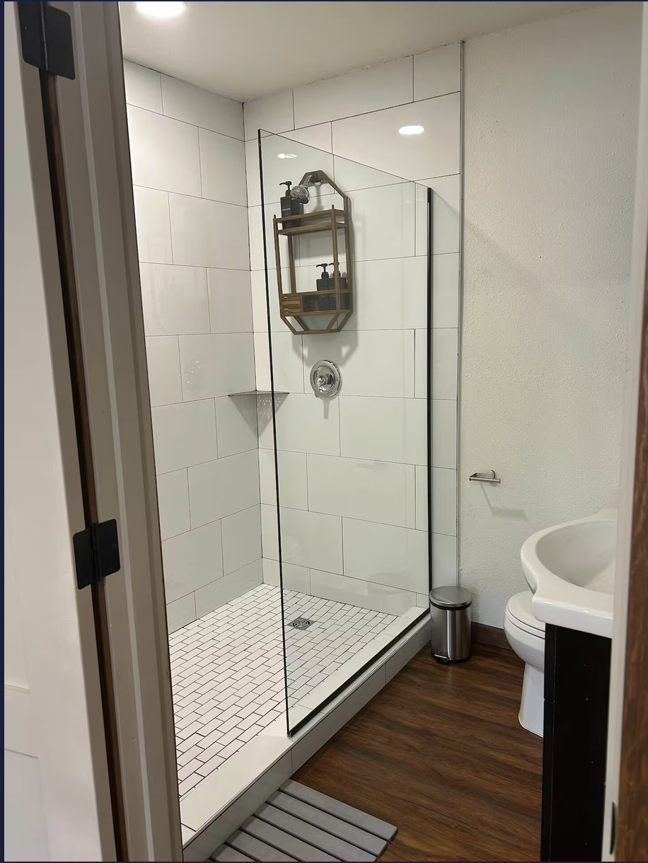 Bathroom shower with glass doors, white tiled walls, and a small corner shelving unit with toiletries; toilet and sink visible outside the shower, wooden floor, and a small trash can.