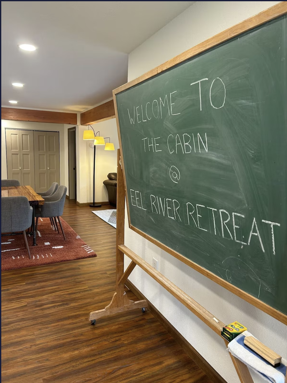 A green chalkboard with the handwritten message, 'Welcome to the Cabin @ Eel River Retreat,' inside a cozy living room with wooden flooring, a dining table with chairs, and a standing lamp with three yellow shades.