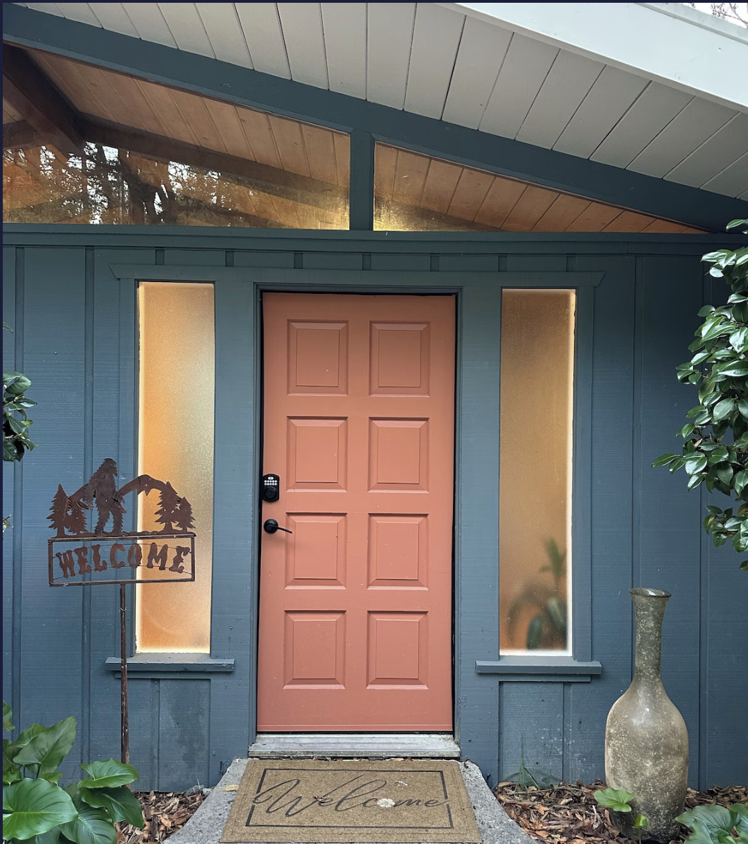 Front door of a house painted in a salmon color, with two glass sidelight windows on each side. A "Welcome" doormat is on the porch, and a decorative metal sign with a bear scene and the word "Welcome" is on the left side. A large vase is on the right, with green plants nearby.