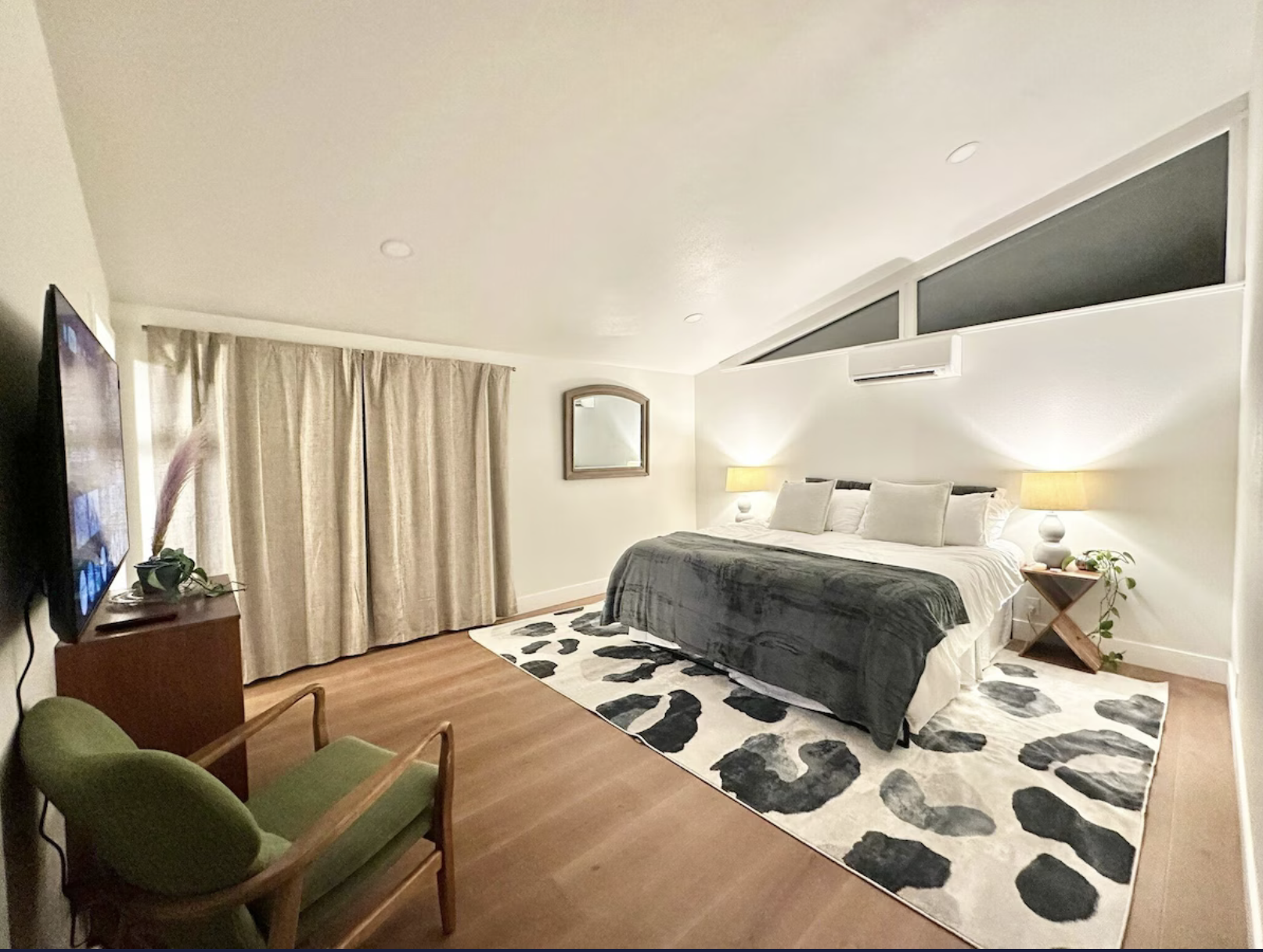A cozy bedroom with a large bed with white and grey bedding, two night lamps, a mirror on the wall, beige curtains, a patterned rug on the wooden floor, and a window with black triangular panes.