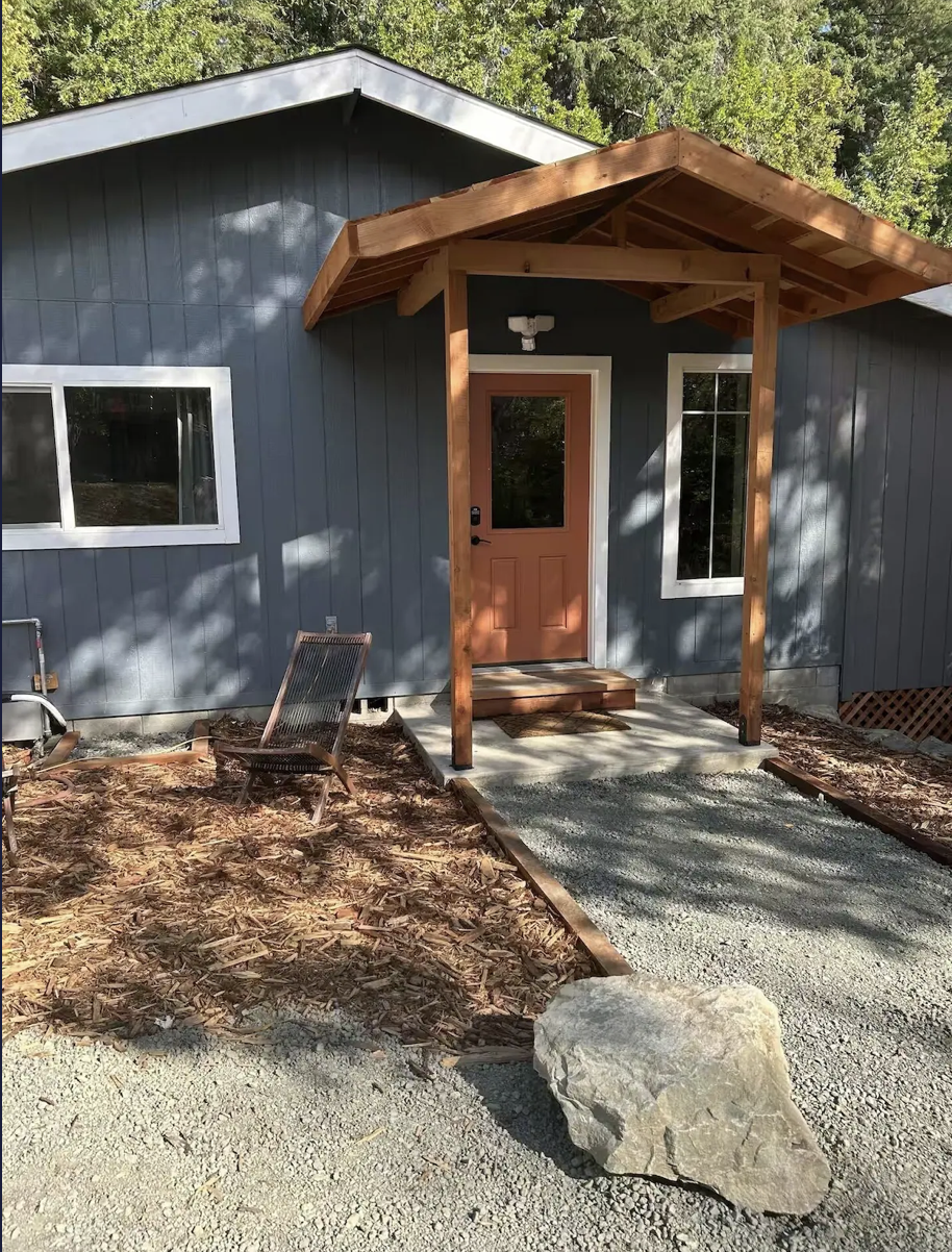 Front porch of a house with a gravel pathway, a large rock, and a wooden chair. The house has a blue exterior with white trim and a small wooden porch roof.