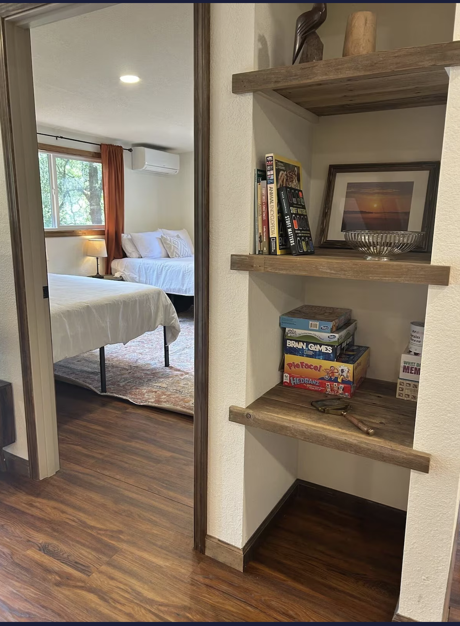 View of a bedroom visible through an open doorway, with two beds and window with orange curtains. Near the doorway, a wooden shelf contains books, framed photo of a sunset, a glass bowl, a small box, a pair of glasses, and a folding knife.