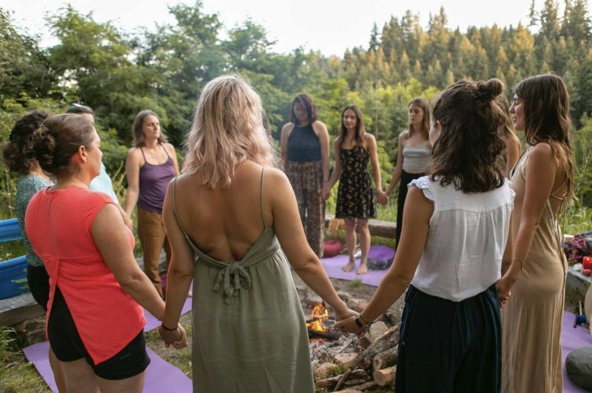 A group of women holding hands in a circle outdoors, participating in a spiritual or meditation ceremony near a campfire, surrounded by trees and nature.