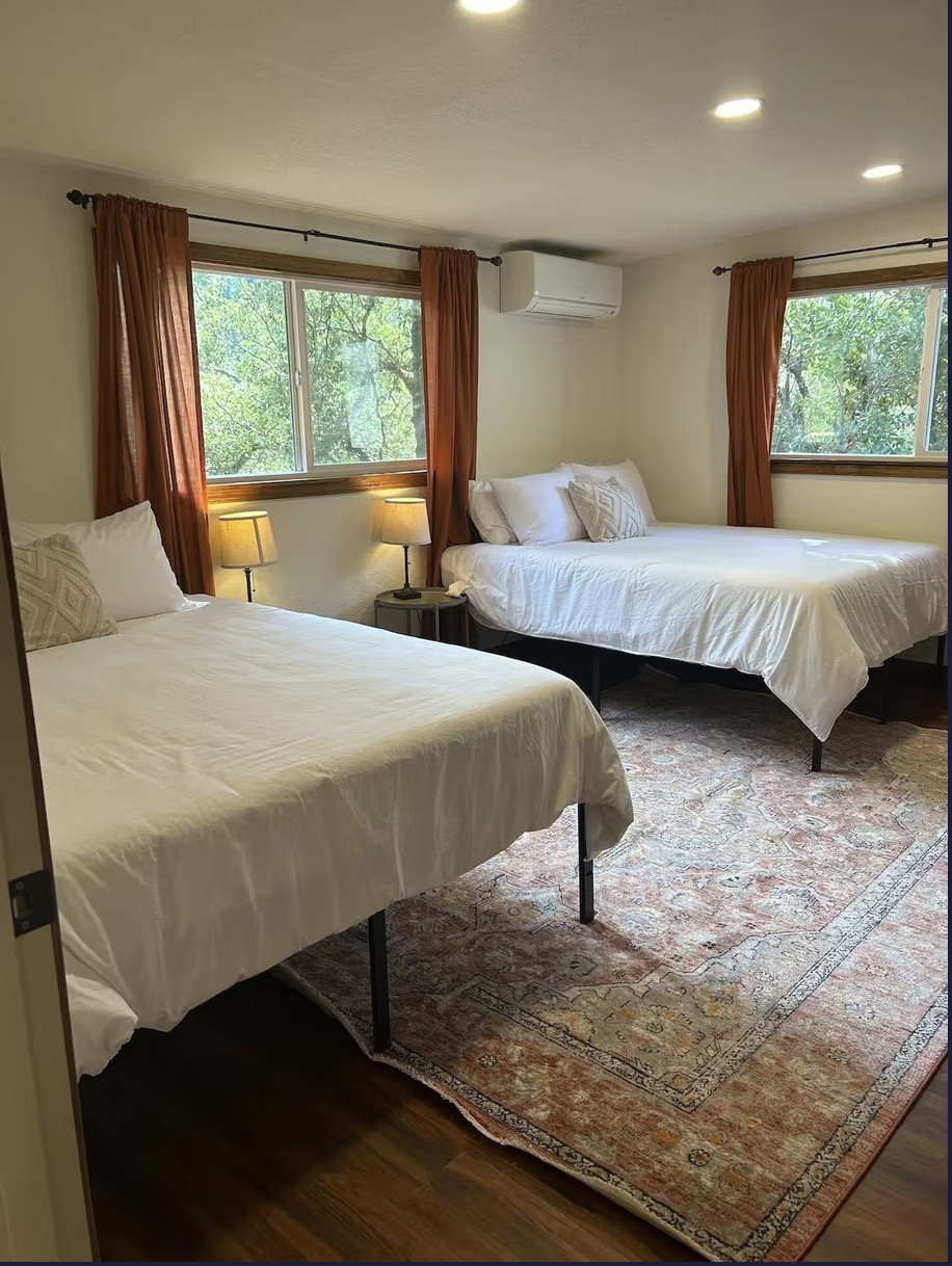 A bedroom with two beds, nightstand with lamp, windows with trees outside, wall air conditioner, and patterned area rug.