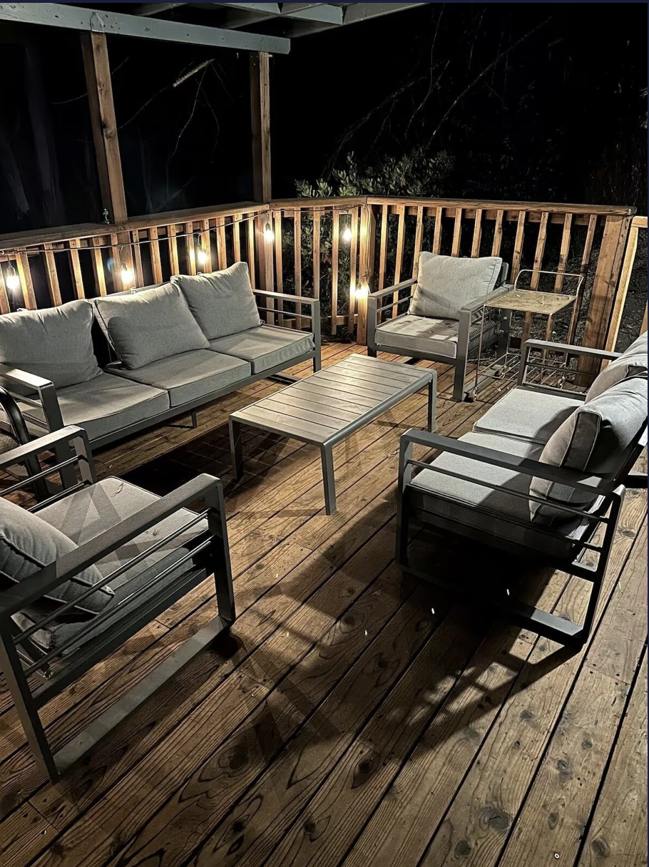 Outdoor wood deck at night with patio furniture including a cushioned sofa and armchairs, metal coffee table, side tables, and string lights on the wooden railing.