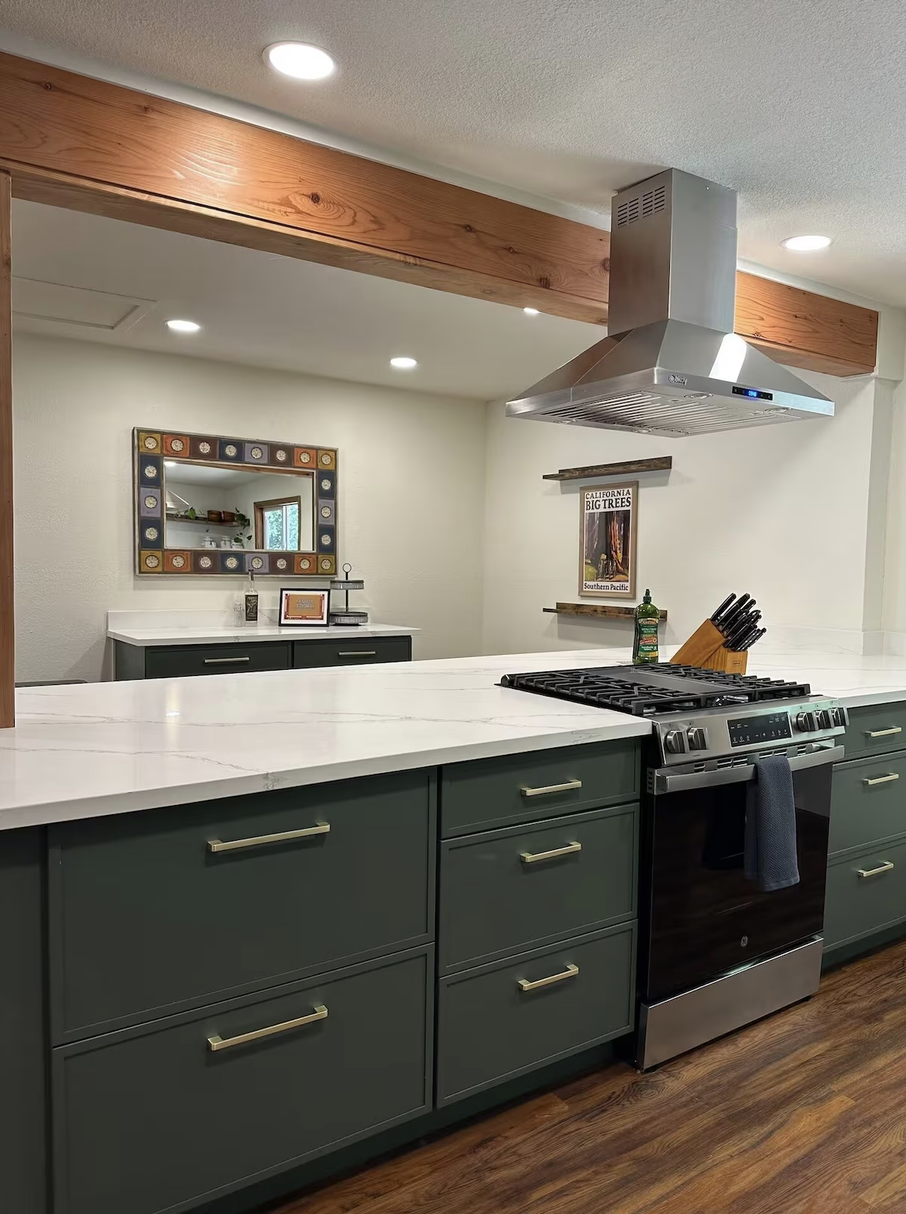 Modern kitchen with green cabinets, white marble countertop, stainless steel oven and range, wooden shelf with a bottle of oil and a knife block, and framed art on the wall.