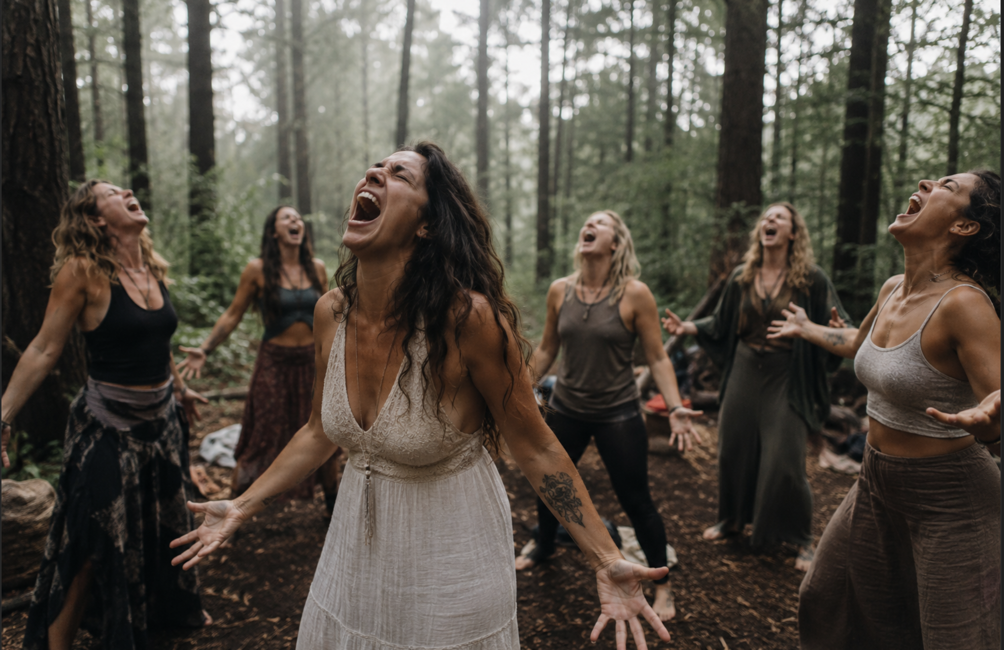 A group of women with long hair, wearing casual, bohemian-style clothing, standing in a circle with eyes closed, while singing or chanting in a forest surrounded by tall trees.