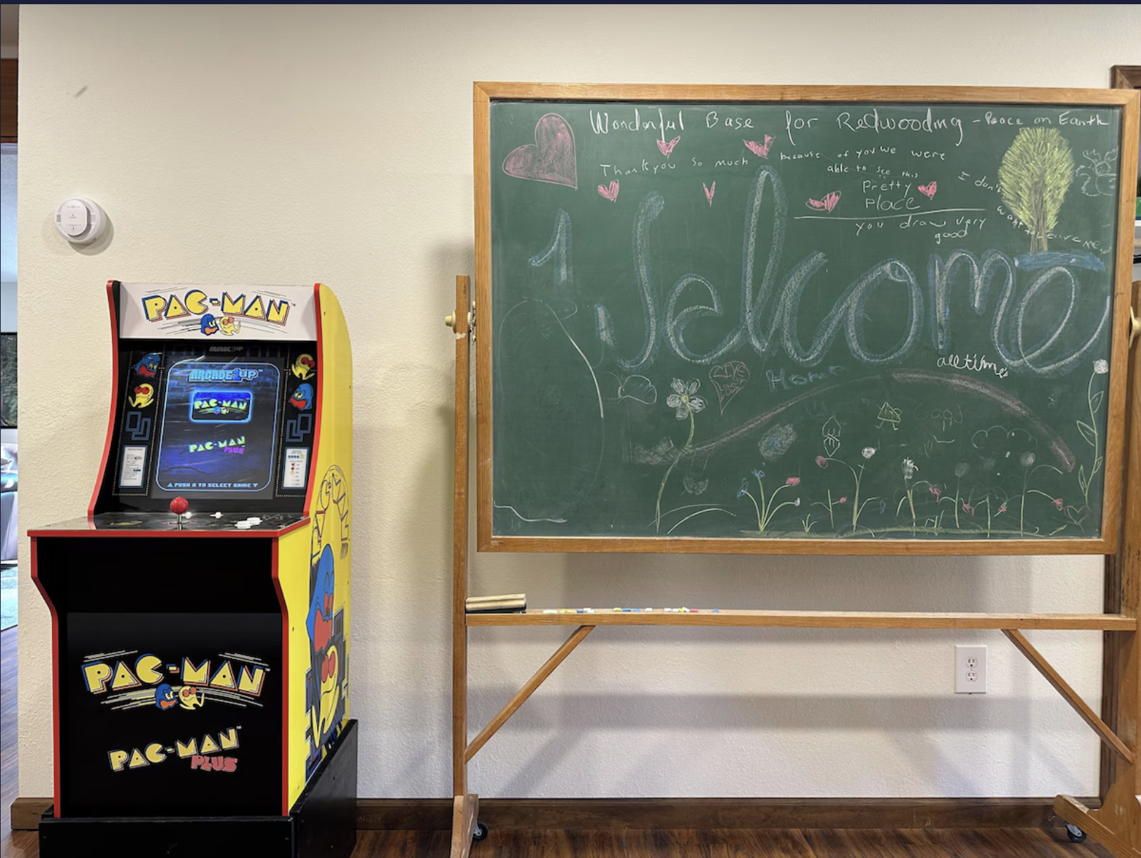 A Pac-Man arcade game machine next to a chalkboard with colorful chalk drawings and writing, including the word 'Welcome' and various doodles, in a room with beige walls and wooden flooring.