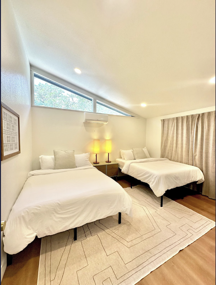 A bedroom with two beds, a beige rug, a nightstand with two lamps, beige curtains, an air conditioning unit, and triangular windows near the ceiling.