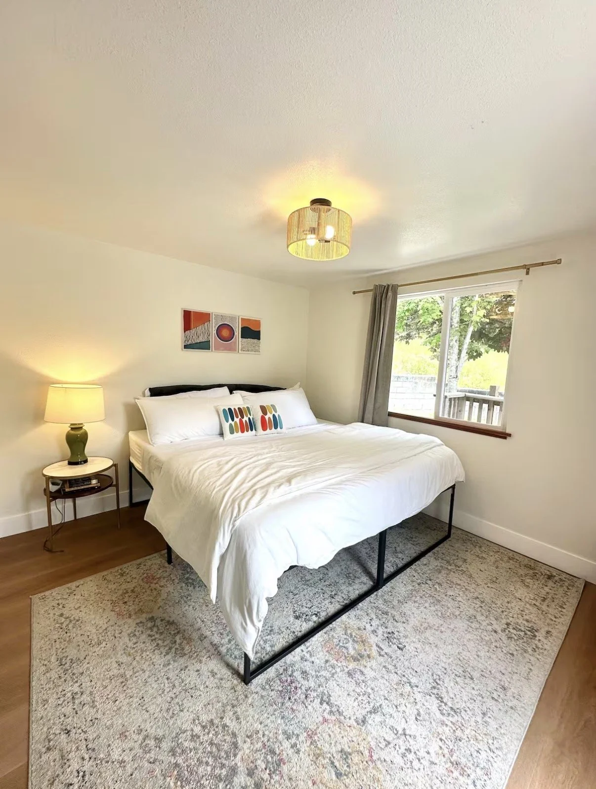 A bedroom with a bed, nightstand, window with curtains, and ceiling light fixture, decorated with artwork and colorful pillows.