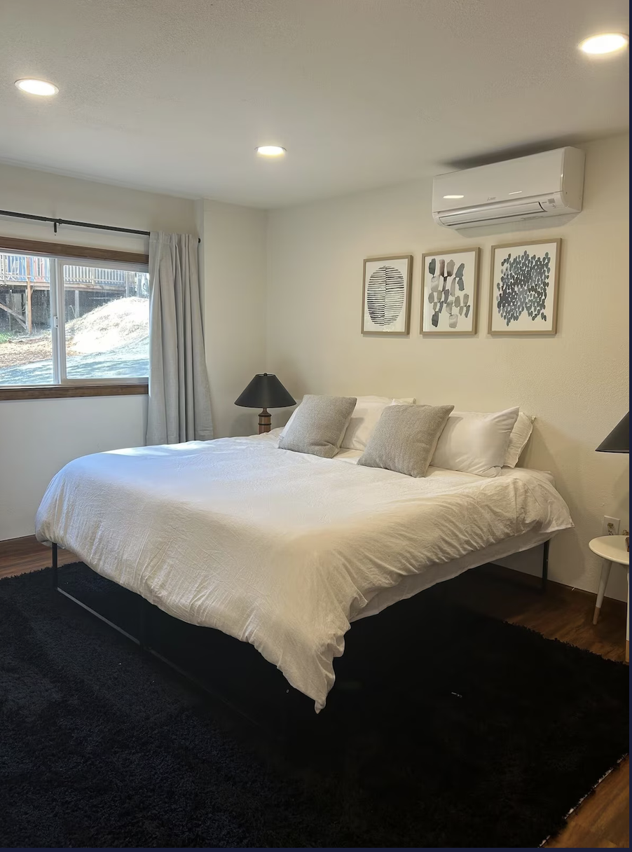 A bedroom with a large bed covered in white bedding, two gray pillows, a black nightstand with a black lamp, a window with curtains, wall art featuring three framed abstract prints, a wall-mounted air conditioner, and a black rug on wooden floor.