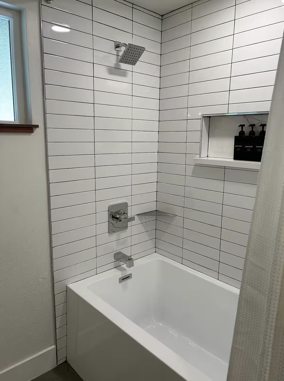 White tiled bathroom shower with square rainfall showerhead, built-in shelf with black bottles, and single-handle faucet above bathtub.