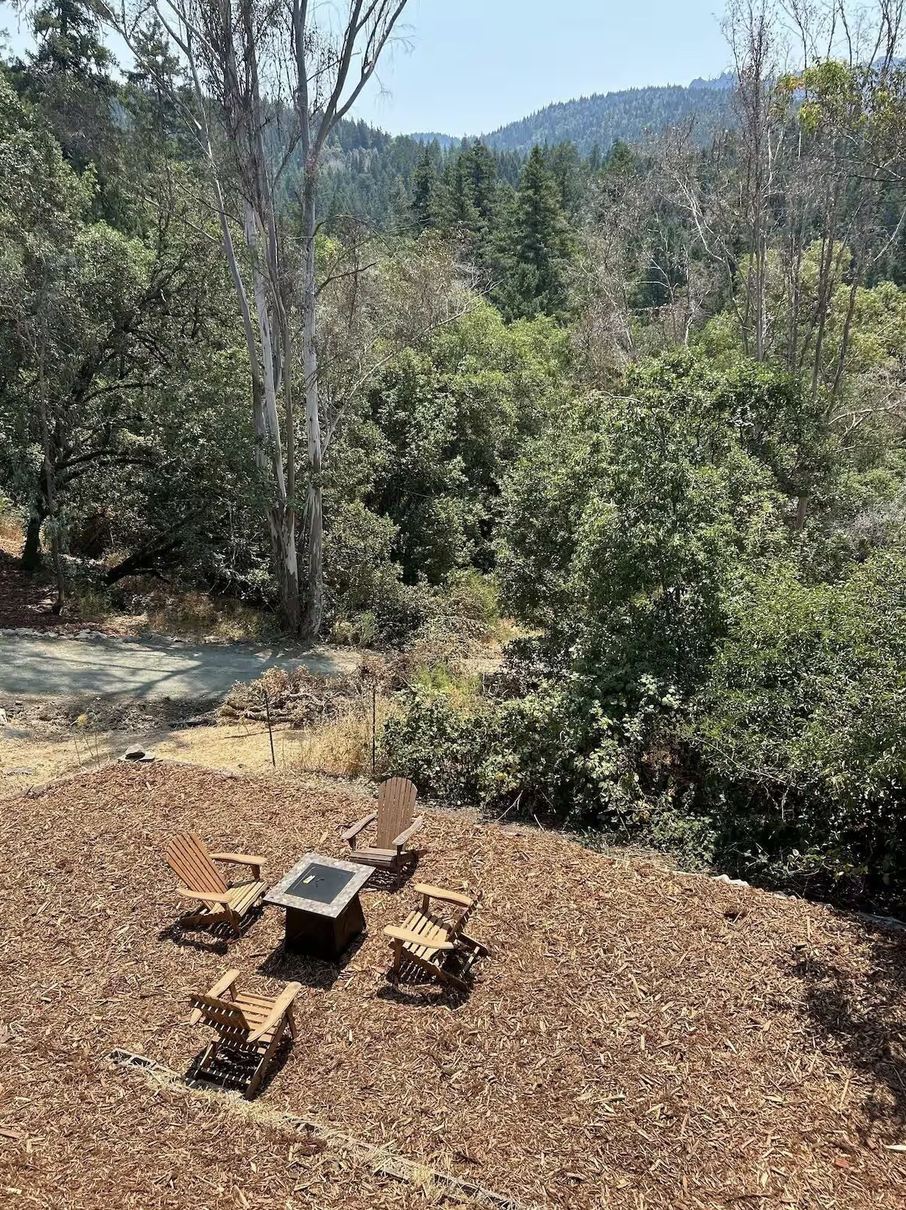 Outdoor seating area with five Adirondack chairs arranged around a fire pit table, on a bed of wood chips, overlooking a forested hillside with tall trees and mountains in the background.