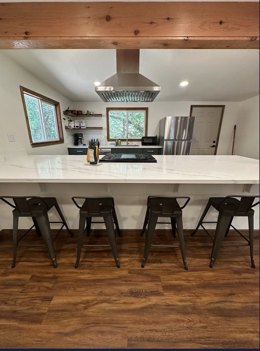 Modern kitchen with white marble island, four black bar stools, stainless steel refrigerator, stainless steel range hood, and wooden floors. Windows show greenery outside.