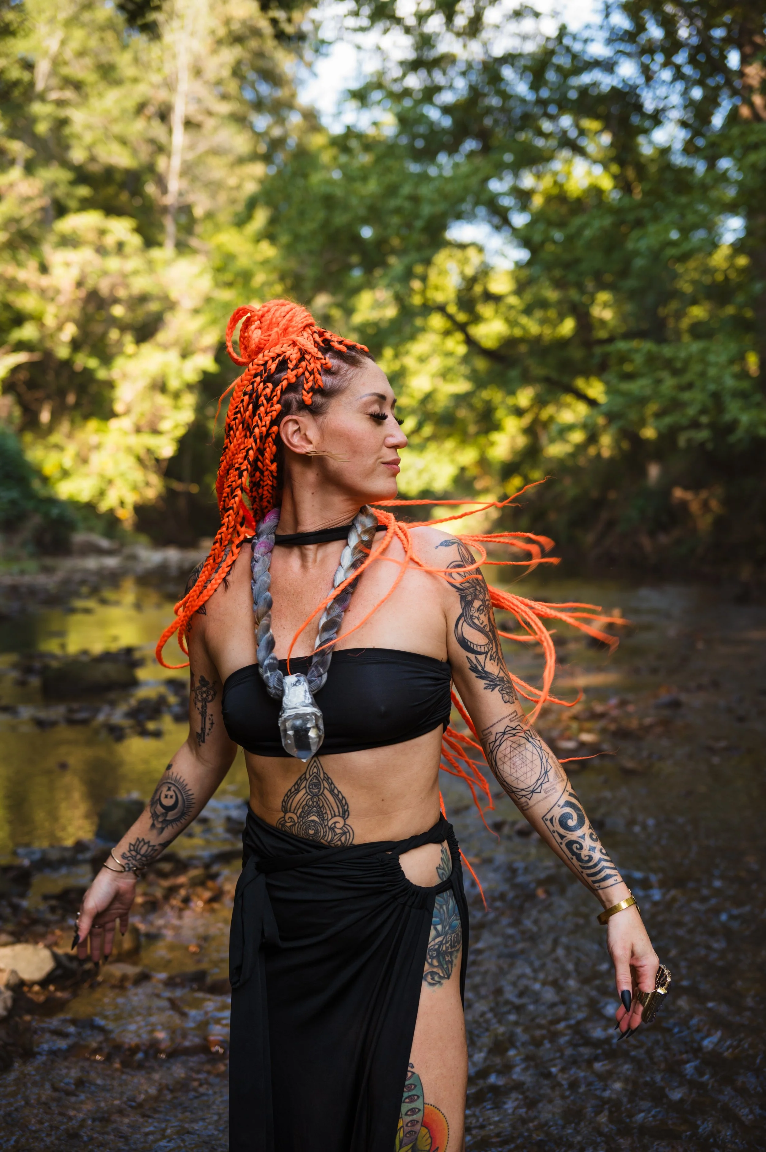 A woman with vibrant orange braids and tattoos on her arms and torso, wearing a black tube top and skirt, standing outdoors in a river with a forest background, looking to her left with her eyes closed.