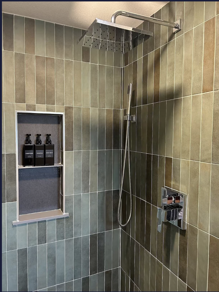 A modern shower with green and brown tiles, a large rainfall showerhead, a handheld showerhead, and a built-in shelf with shampoo and soap.