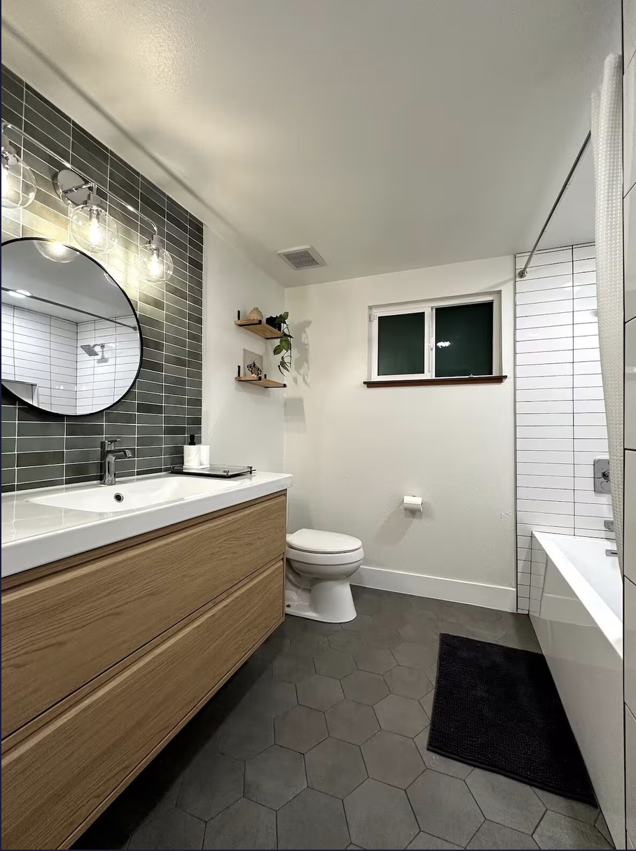 Modern bathroom with a wooden vanity, large mirror, gray subway tile wall, and hexagonal gray floor tiles. Features a toilet, small window, and a bathtub with a white shower curtain.
