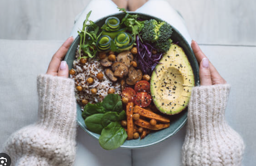 A person wearing a cozy cream-colored sweater holding a bowl of colorful vegetarian salad with various vegetables and toppings.